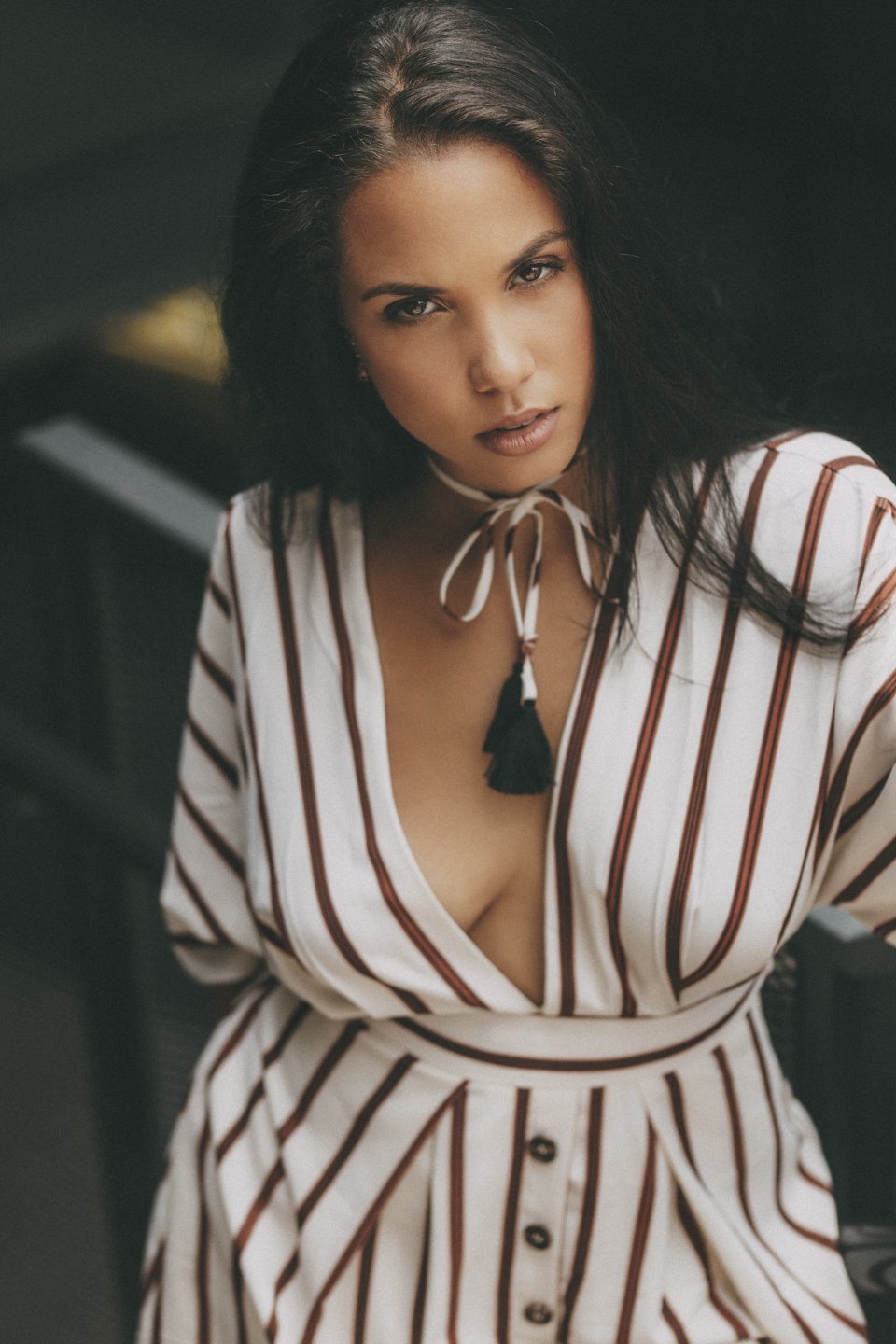 A woman with dark hair and light skin wearing a striped deep V-neck blouse with buttons and a black tassel necklace, looking directly at the camera.