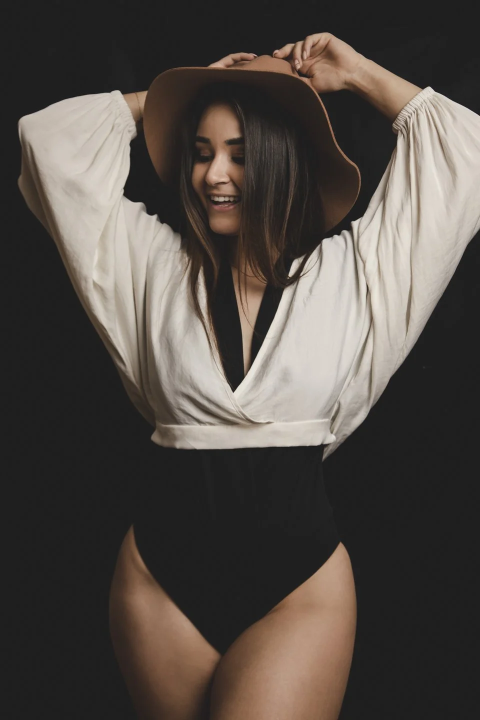 Young woman smiling, wearing a wide-brimmed hat, white top, and black bottoms, standing against a black background.