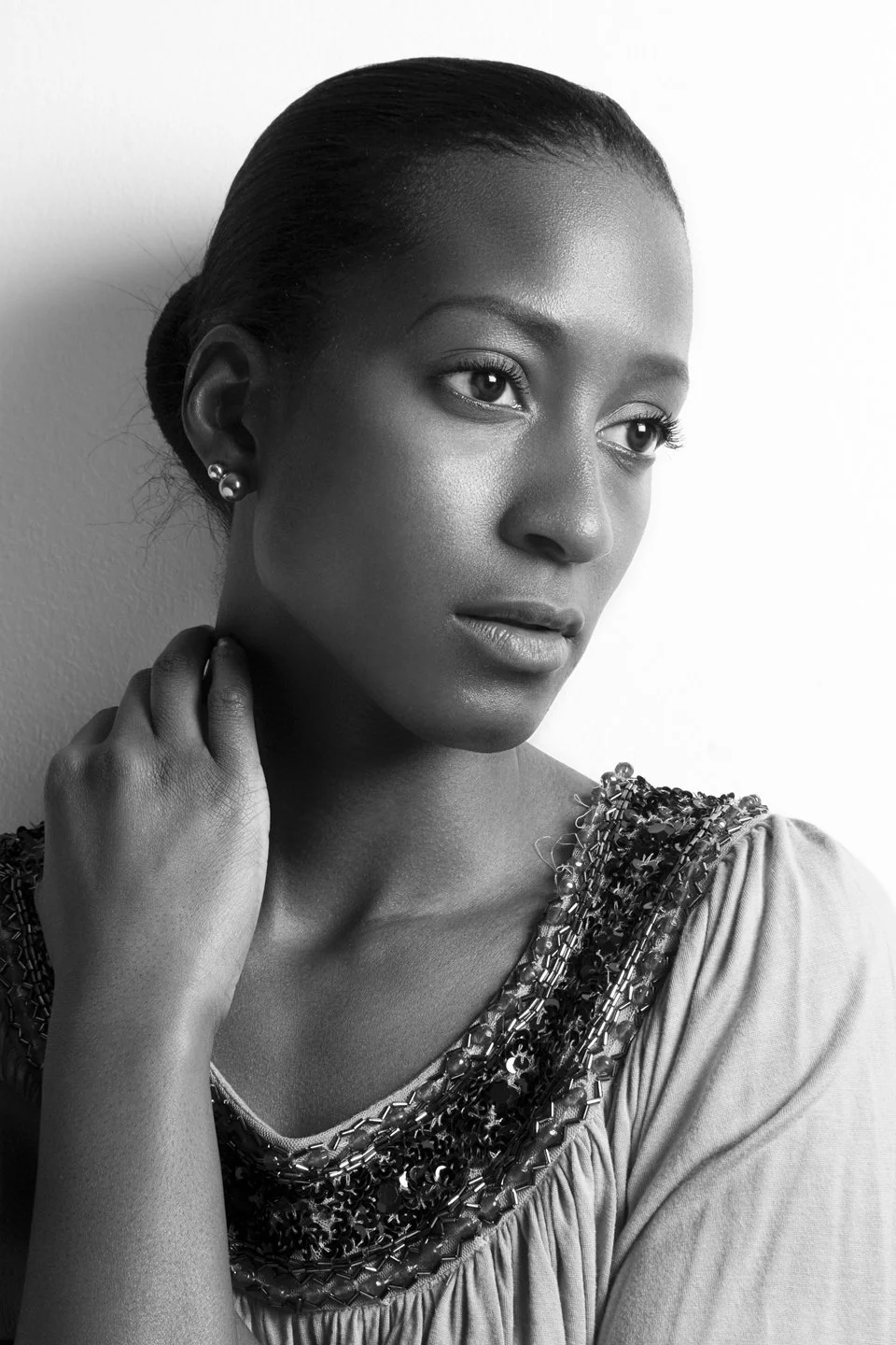 Black and white portrait of a woman with smooth skin and a serious expression, her hair slicked back, wearing pearl earrings and an embellished garment with beading and sequins.