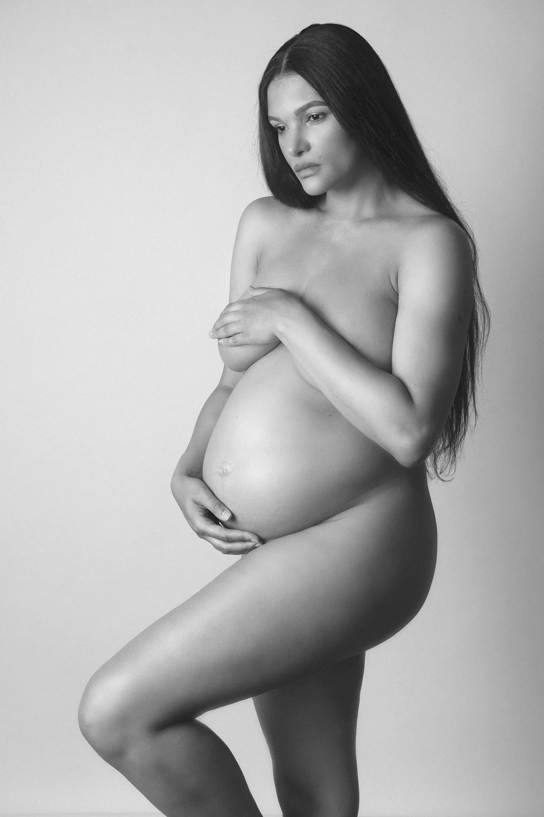 Black and white portrait of a pregnant woman with long hair, standing and holding her belly.
