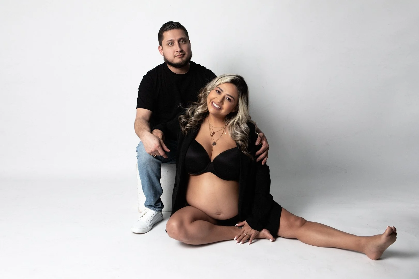 Couple posing for a maternity photo shoot on a white background, with the expecting mother sitting on the floor and the father sitting behind her.