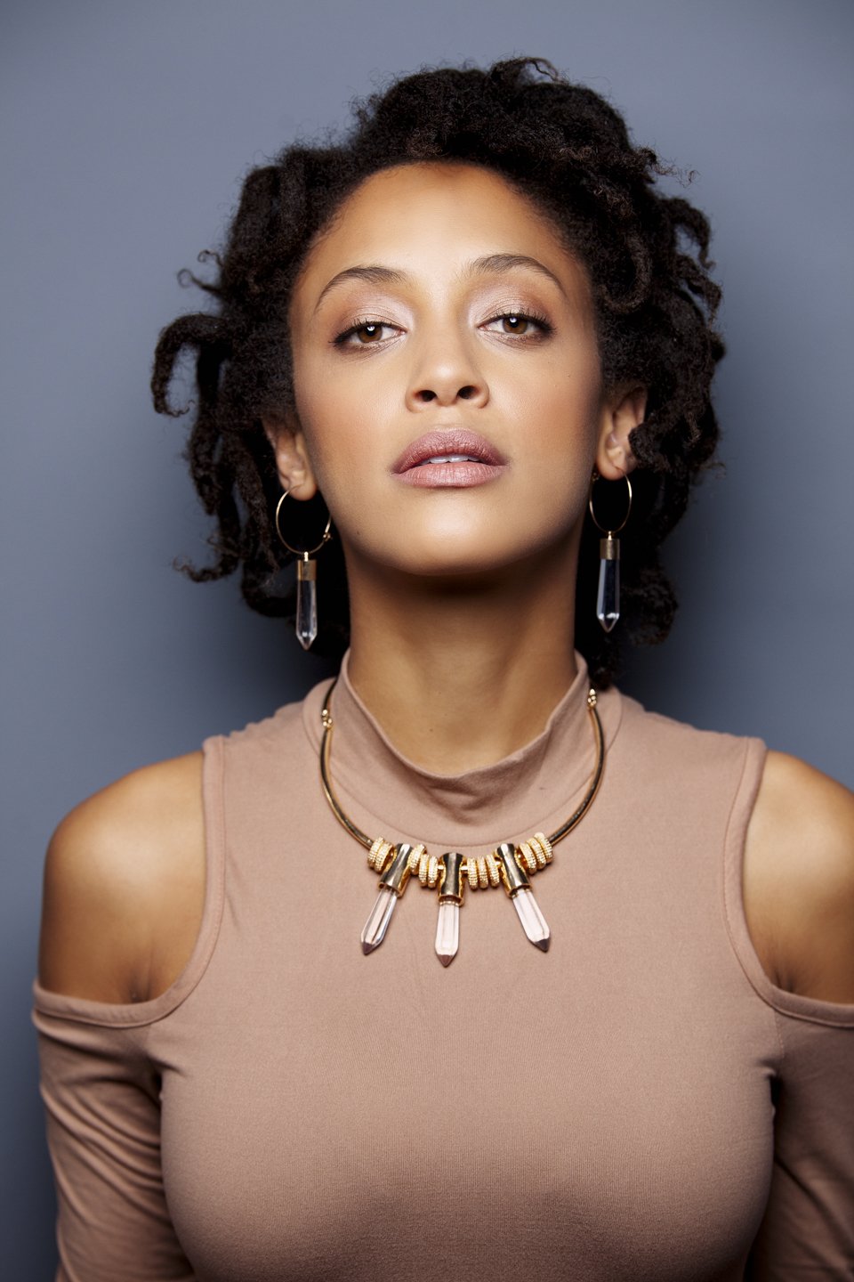 A woman with short, curly hair and hoop earrings, wearing a beige top with a cold shoulder design and a statement necklace with crystals, posing against a dark background.