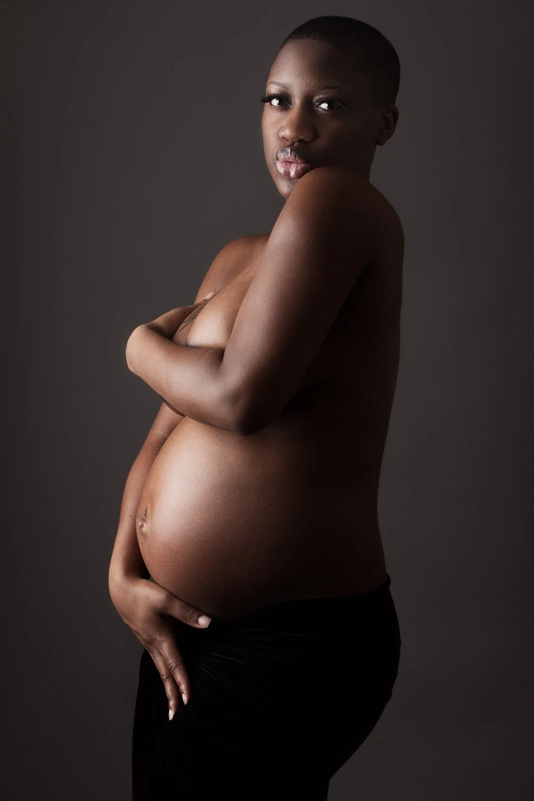 Pregnant woman posing with arms crossed over chest in dark background.