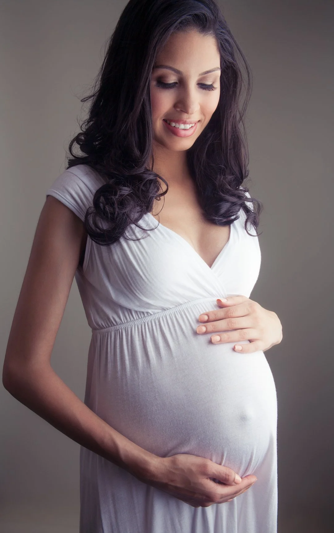 Pregnant woman in a white dress smiling and holding her belly.