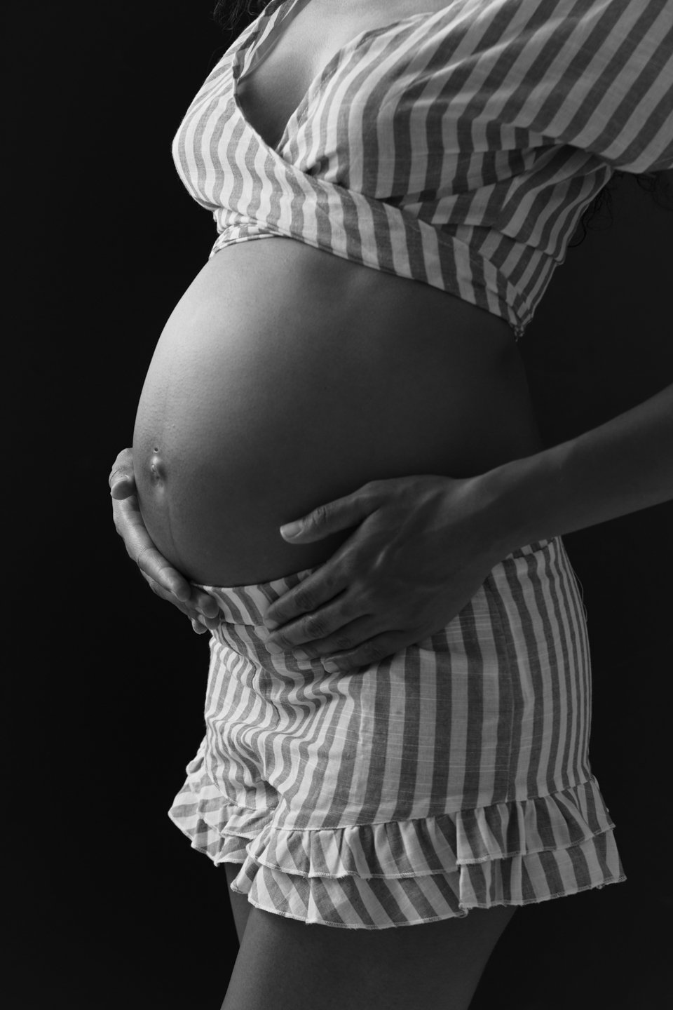 Black and white photo of a pregnant woman cradling her belly, wearing striped pajamas and a crop top.