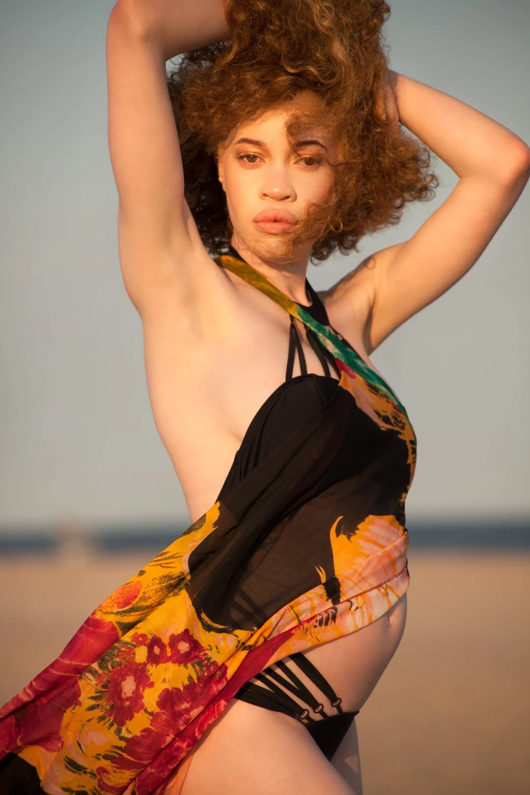 A person posing on a beach wearing a colorful, patterned wrap over a black swimsuit, with arms raised and hair blowing in the wind. The background features a clear sky and sandy beach.