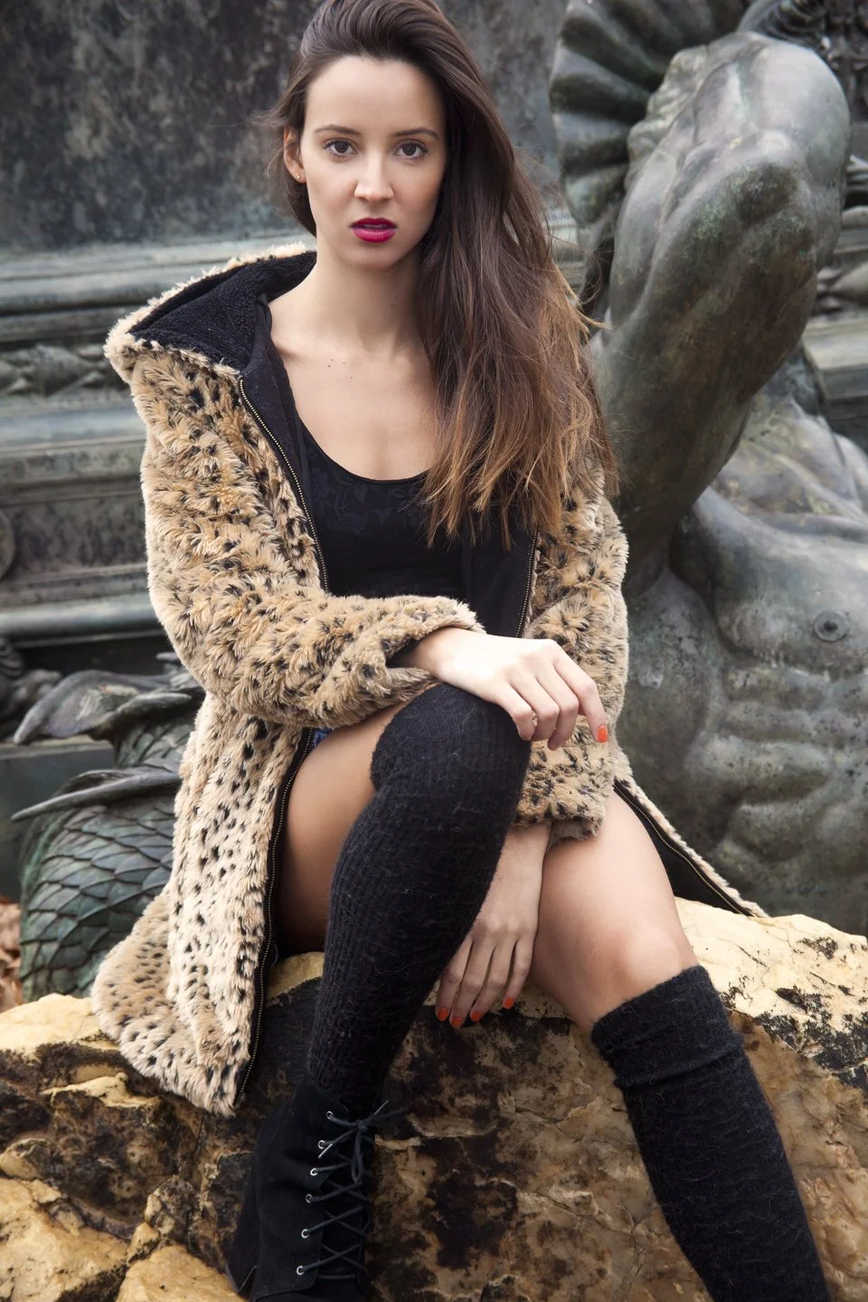 A woman with long brown hair and red lipstick sitting on a rock in front of a large sculpture of a face, wearing a leopard print coat, black top, black knee-high socks, and black boots.