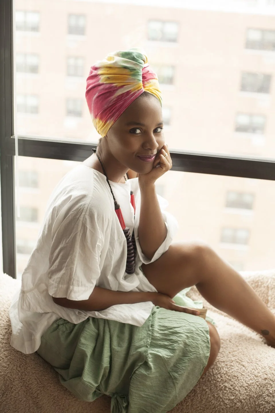 A woman sitting on a bed near a window, wearing a colorful headwrap, a white shirt, and light green skirt, looking at the camera with her head resting on her hand.