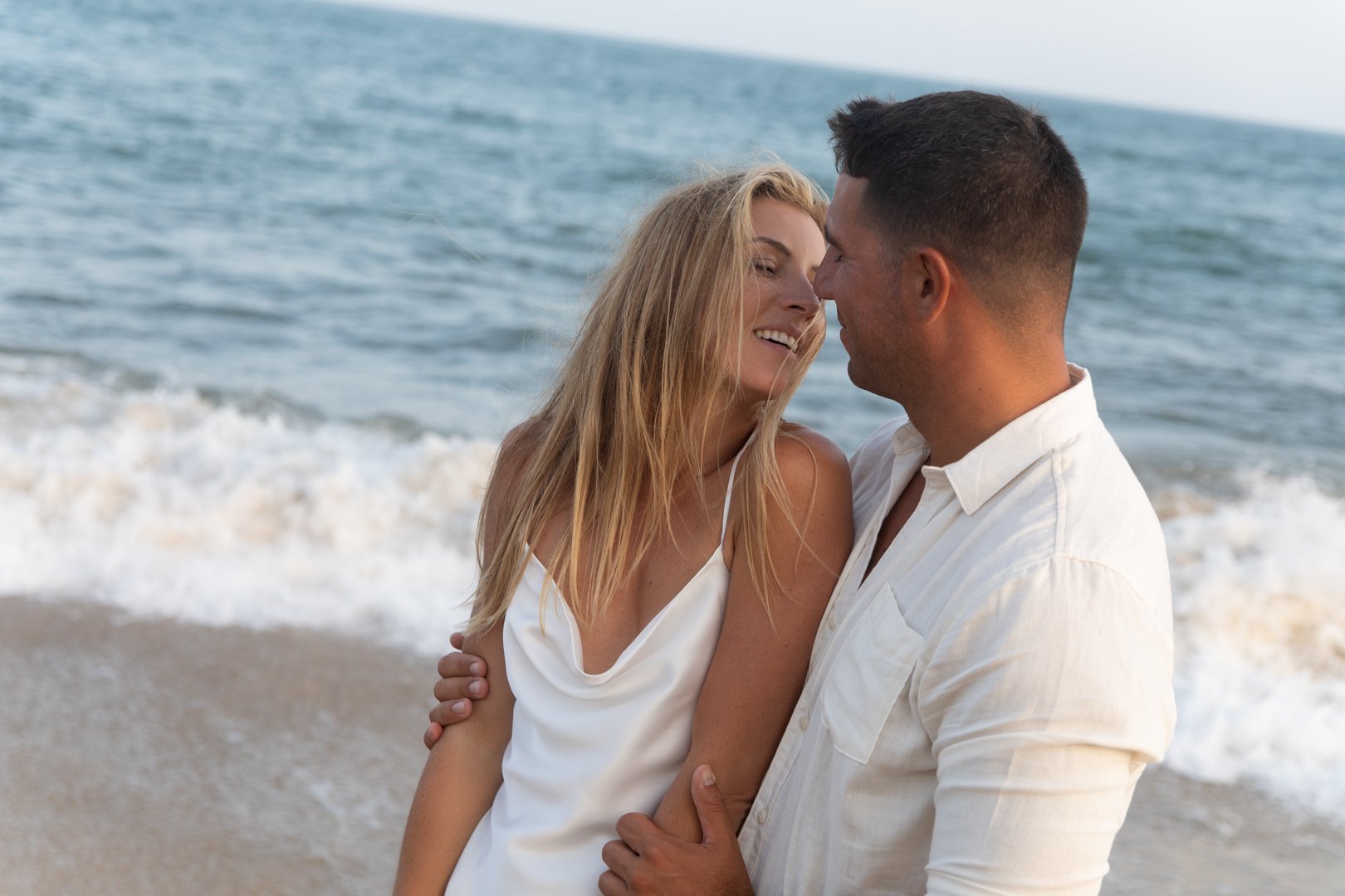 A couple embracing and smiling at each other on the beach near the ocean.