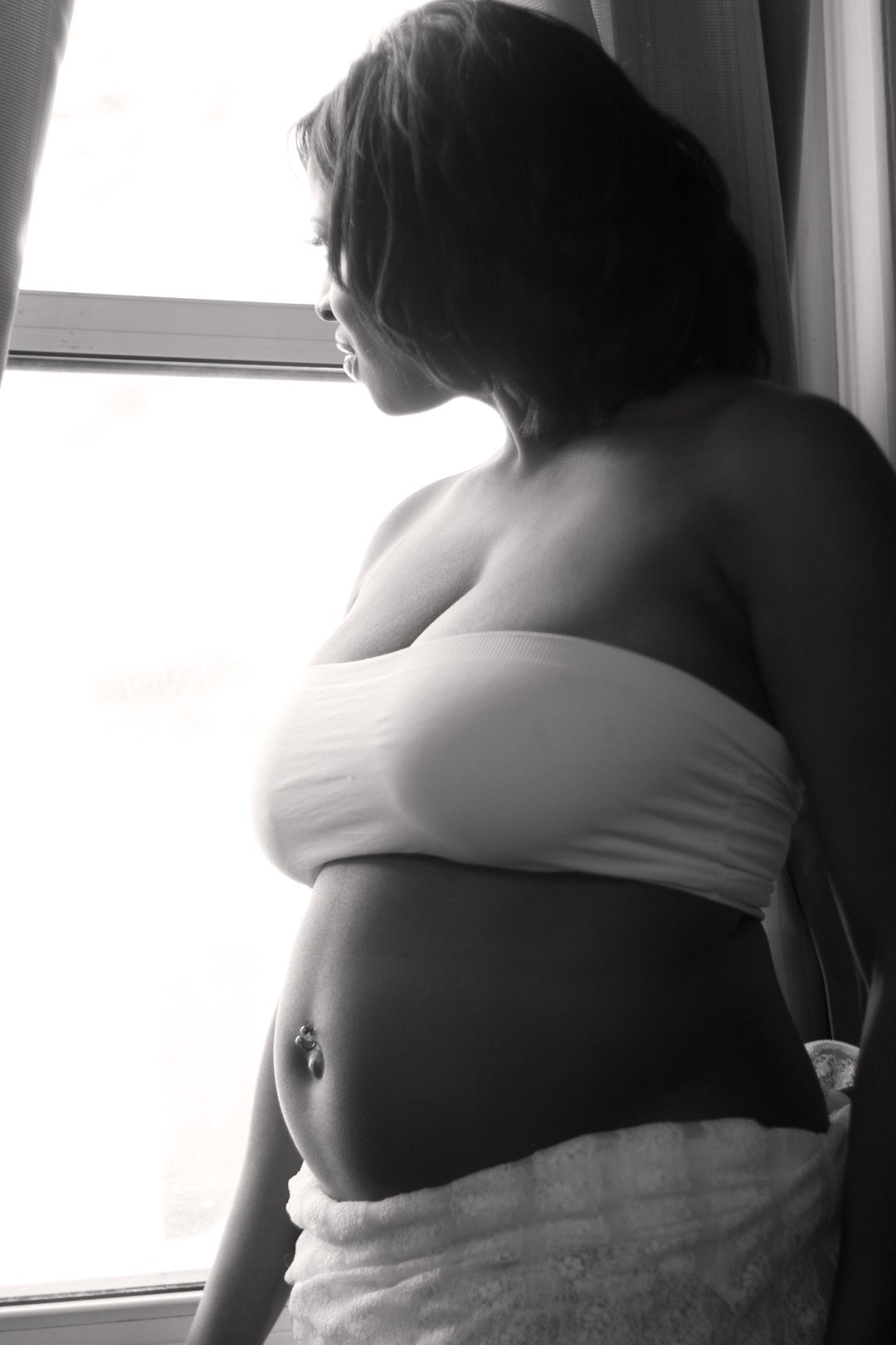 Black and white photo of a pregnant woman wearing a bandeau top, looking out of a window.