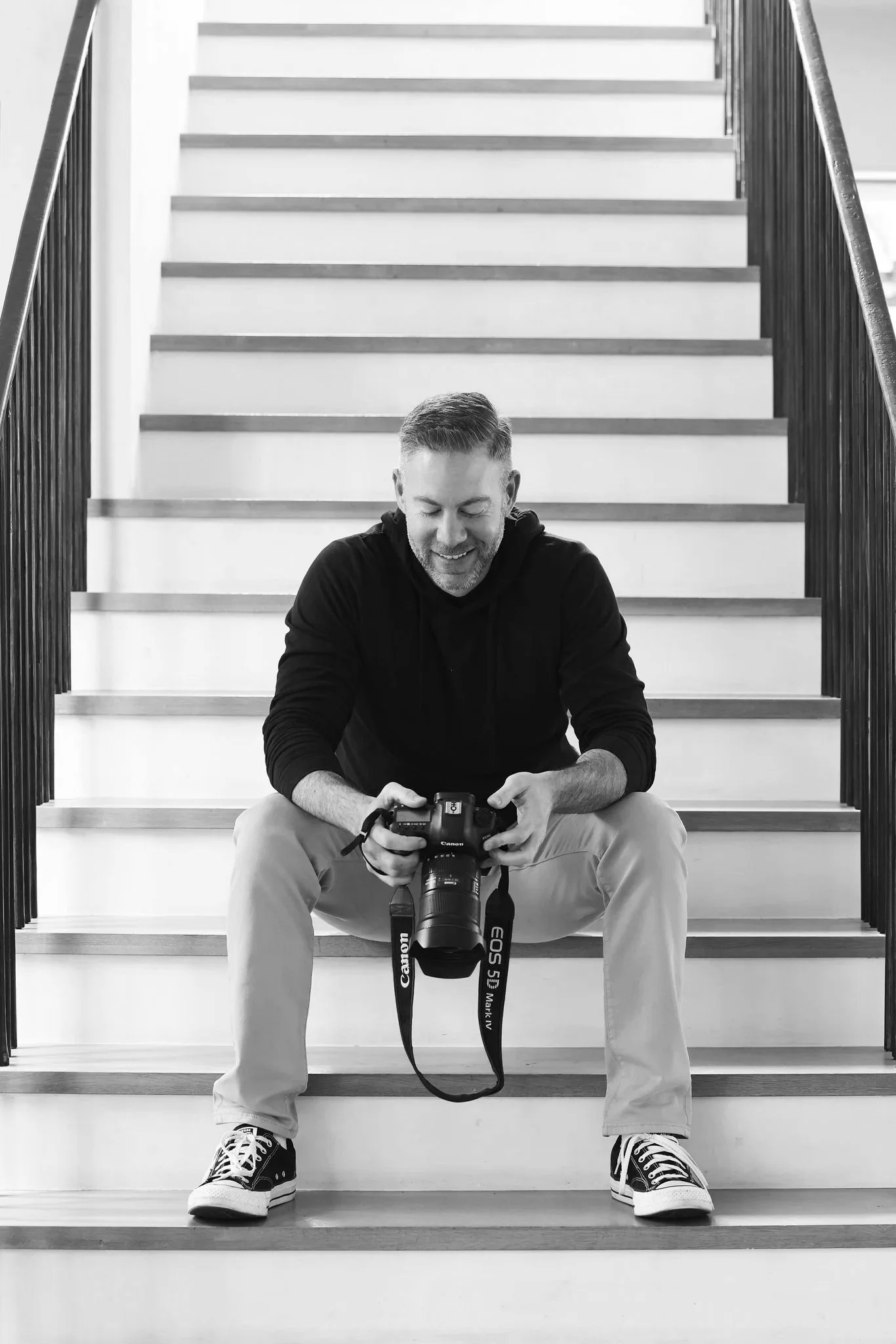A smiling man with a beard sitting on stairs, looking at a camera in his hands. He is wearing a black hoodie, beige pants, and sneakers. The stairs are wooden with dark railings on each side. The photo is in black and white.