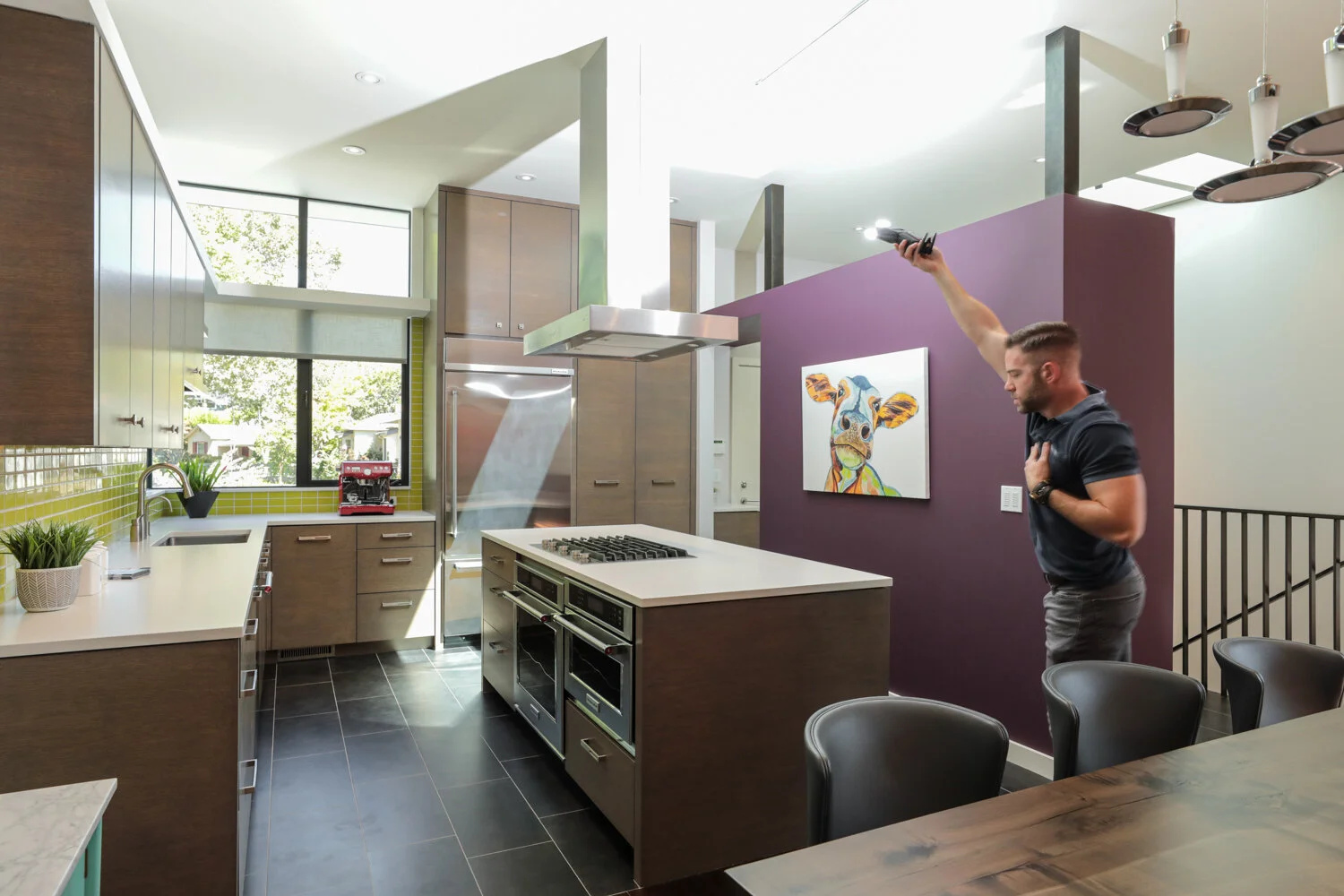 A man holding a remote control and an appliance, standing in a modern kitchen with purple accent wall and colorful cow painting.