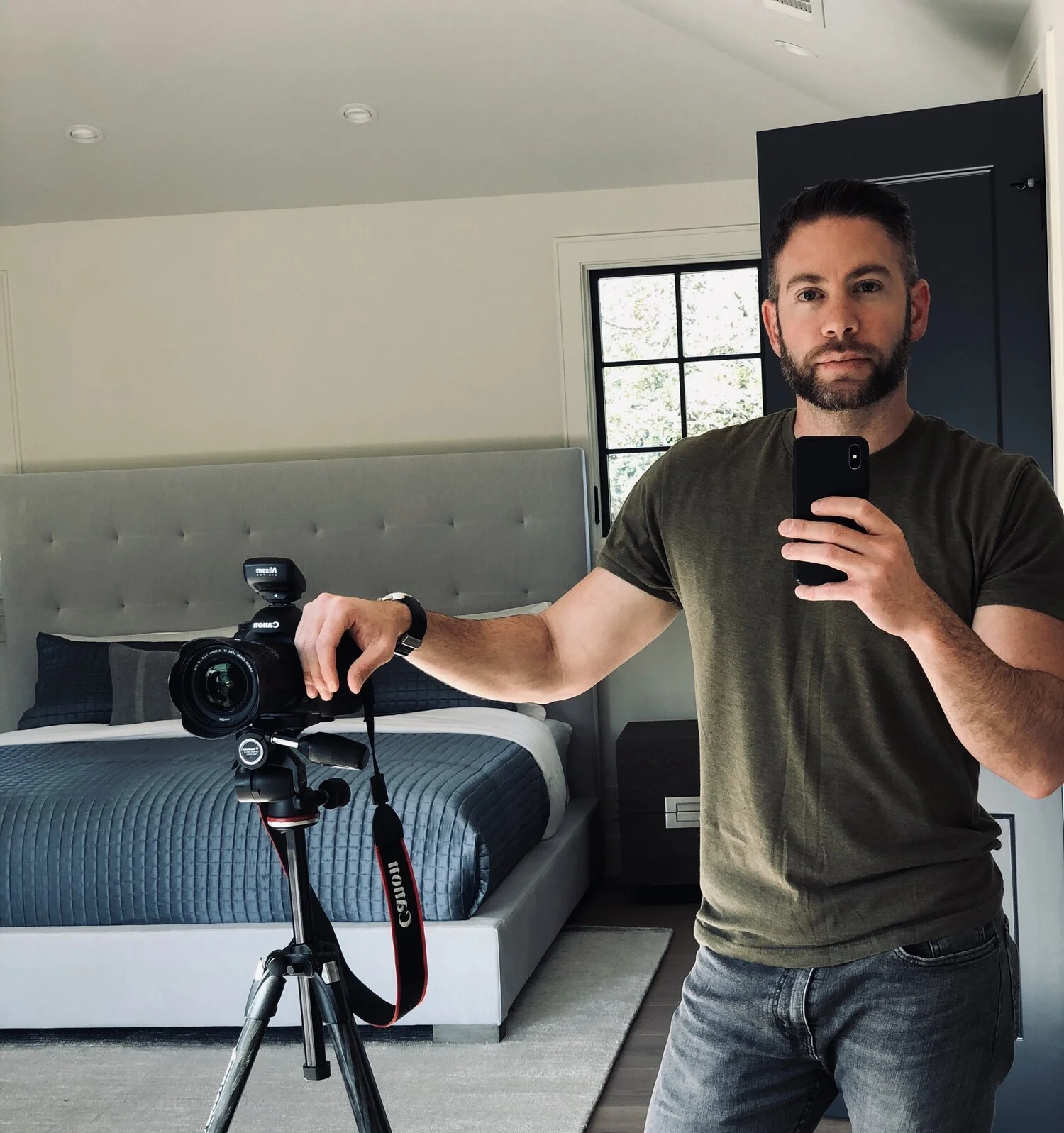 A man taking a mirror selfie in a bedroom, holding a camera with a tripod, and standing next to a bed with a gray quilted bedspread. He has dark hair, a beard, and is wearing a dark green T-shirt and gray jeans. There is a window with black framing a