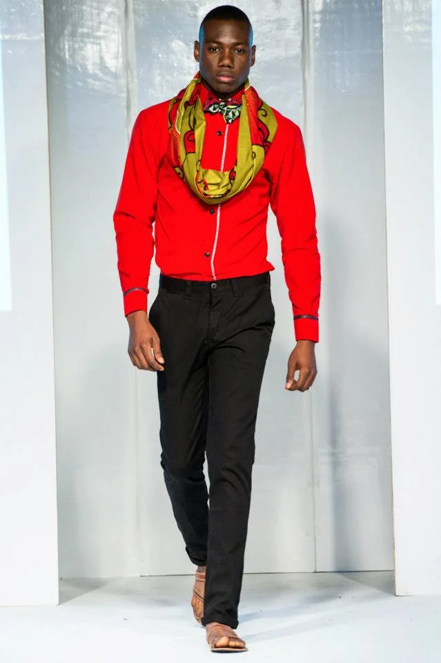 A male model walking on a runway, wearing a red shirt, black pants, tan sandals, and a colorful scarf around his neck.