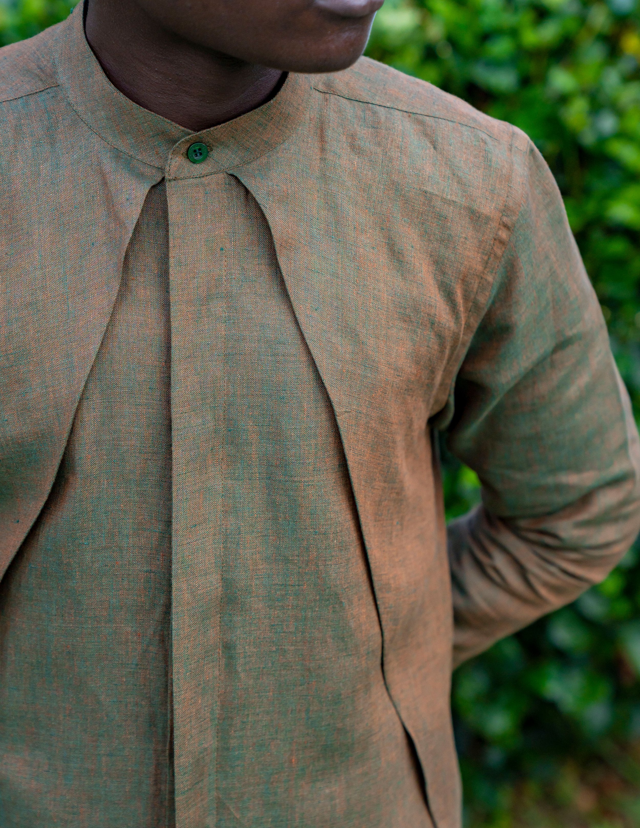 Close-up of a person wearing a brownish-gray button-up shirt with a yellowish tinge, standing outdoors with blurred green foliage in the background. The person's face is partially visible, with the focus on the upper torso and neck.