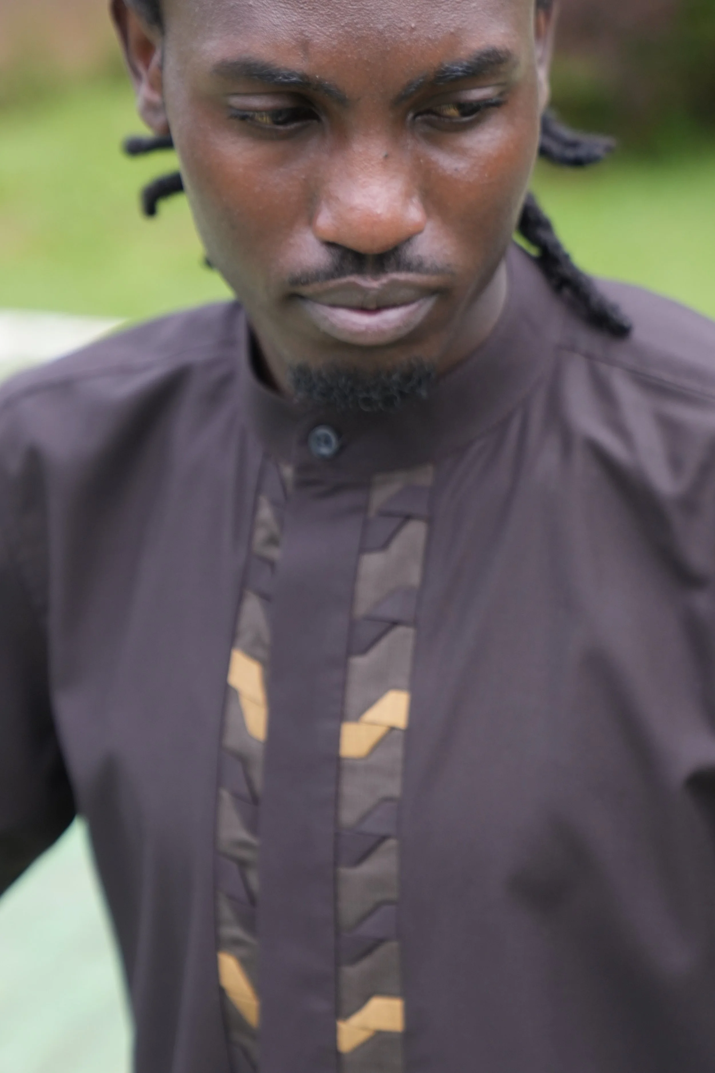 Close-up of a man with medium brown skin, dreadlocked hair, wearing a dark shirt, looking downward with a serious expression.