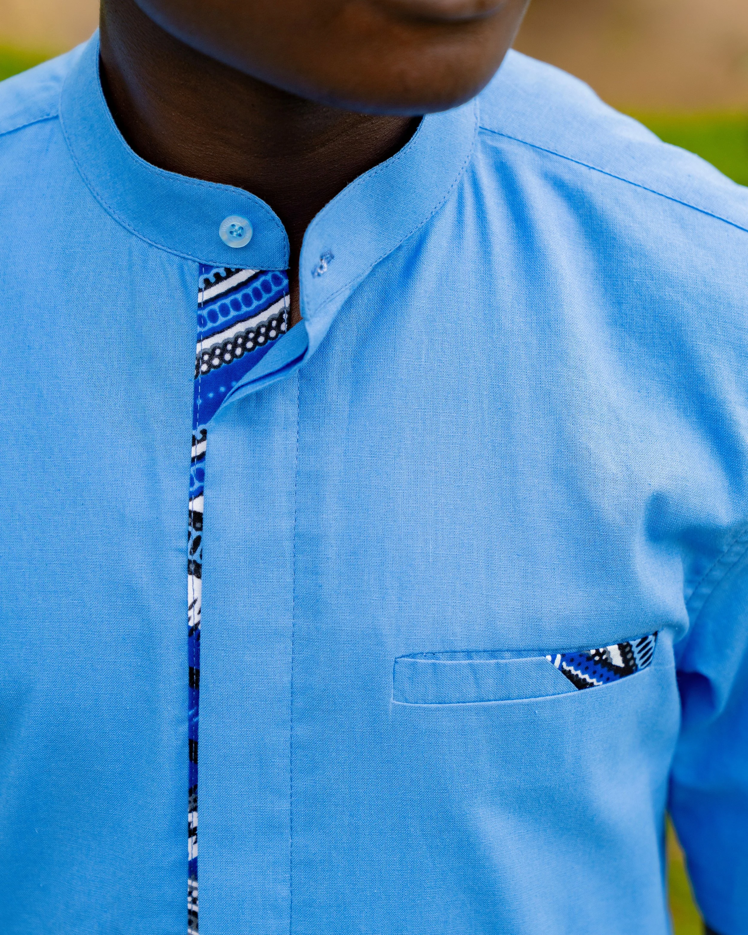 Close-up of a man wearing a light blue shirt, featuring a patterned inner collar and a pocket with a matching patterned trim, positioned outdoors.