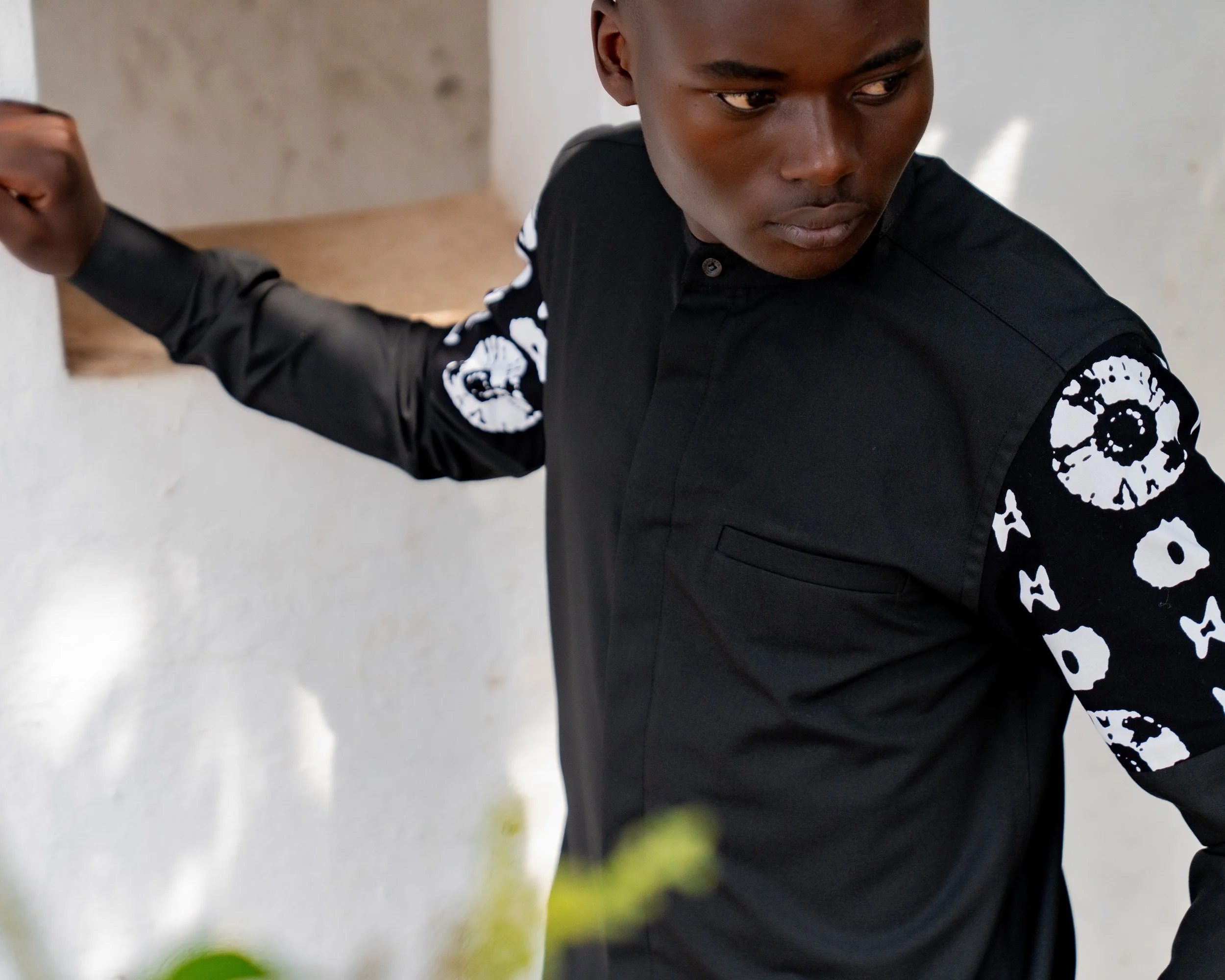 A young man with dark skin and short hair, wearing a black shirt with white skull designs on the sleeves, is seen leaning against a white wall, looking downward with a serious expression.