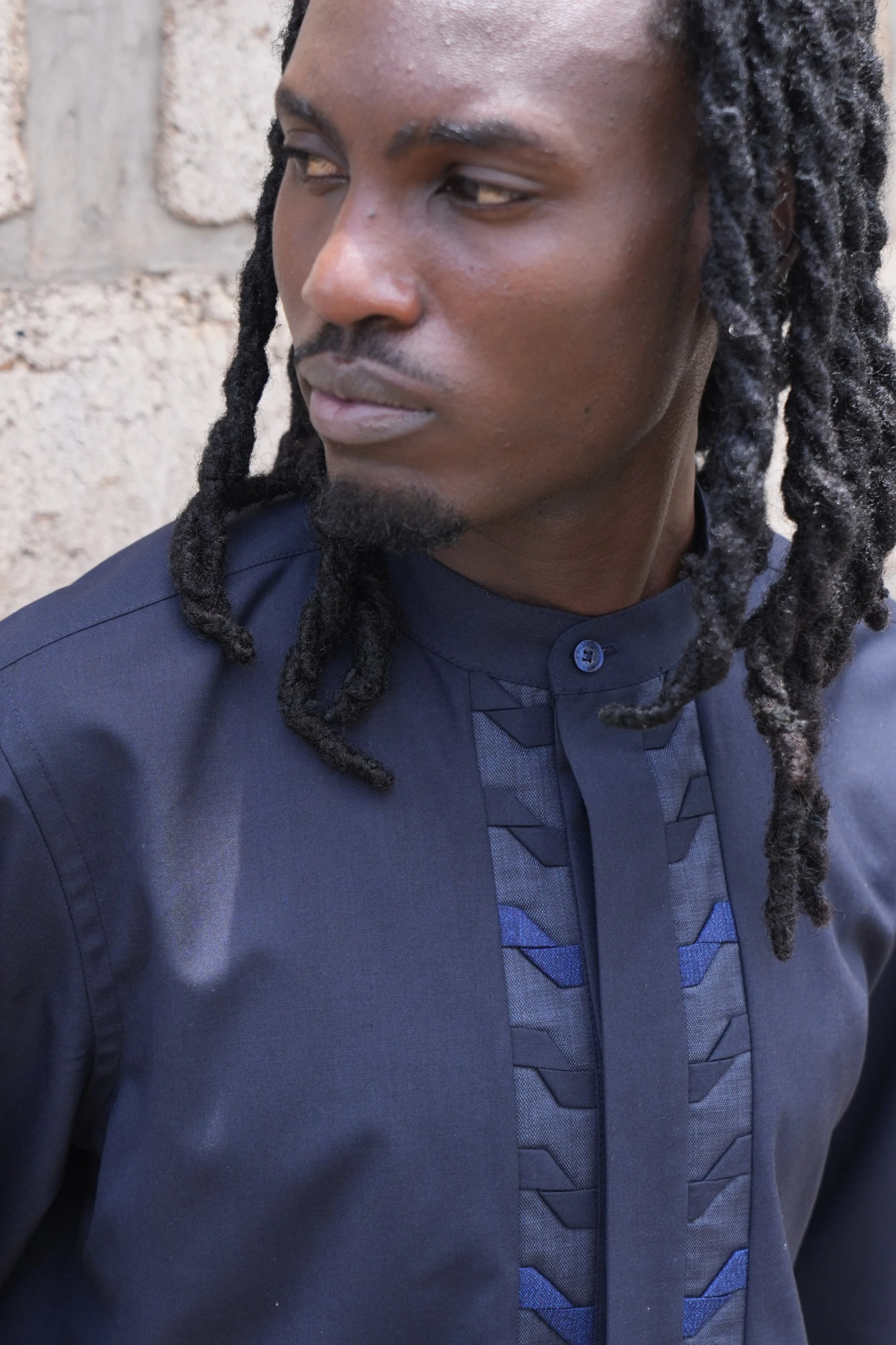 A man with long dreadlocks, wearing a dark blue/black traditional African shirt with geometric embroidery, standing against a stone wall.