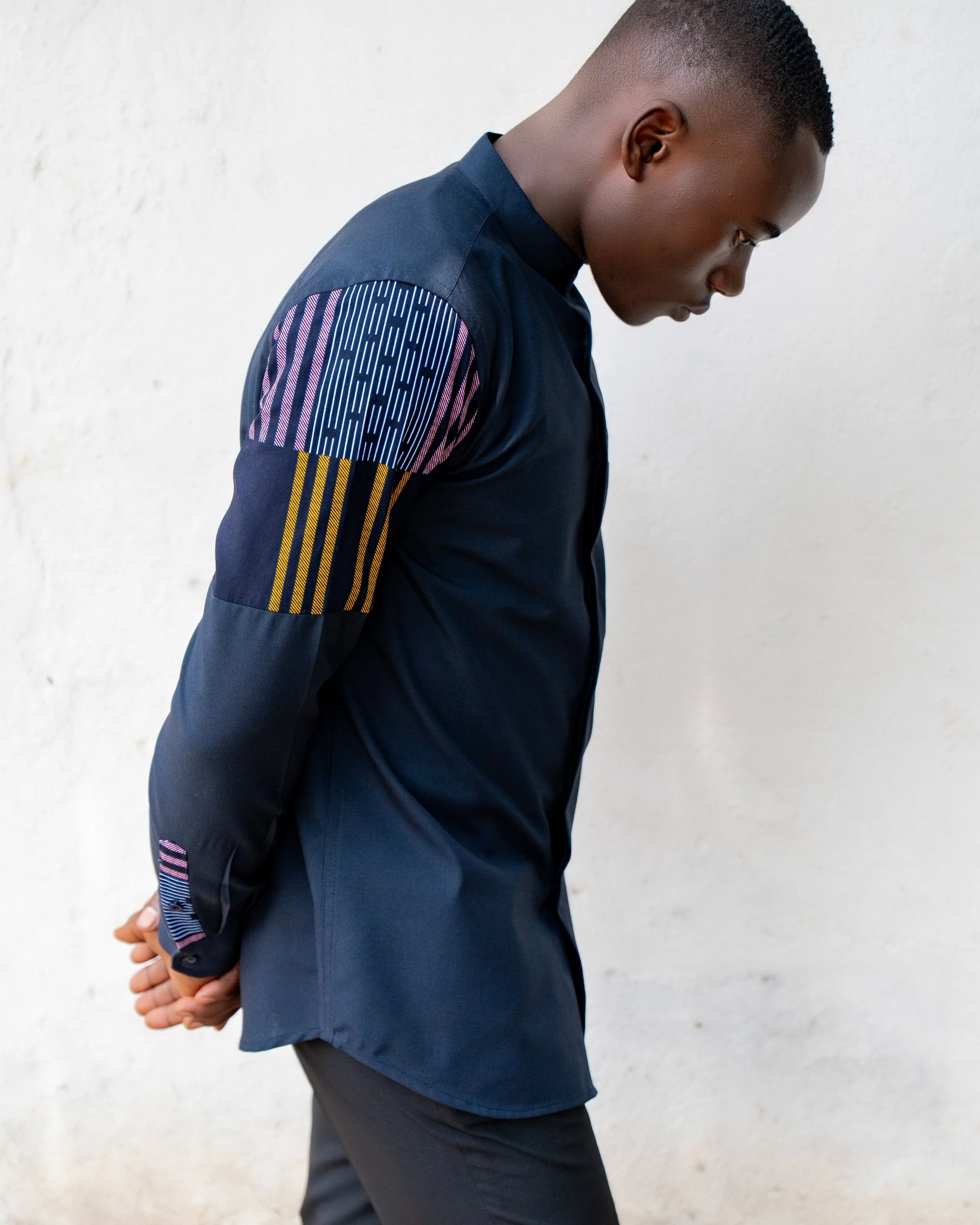 A young man with dark skin and short curly hair, dressed in a navy jacket with colorful patterned accents on the sleeves, standing against a plain white wall, with his hands clasped behind his back, looking down.