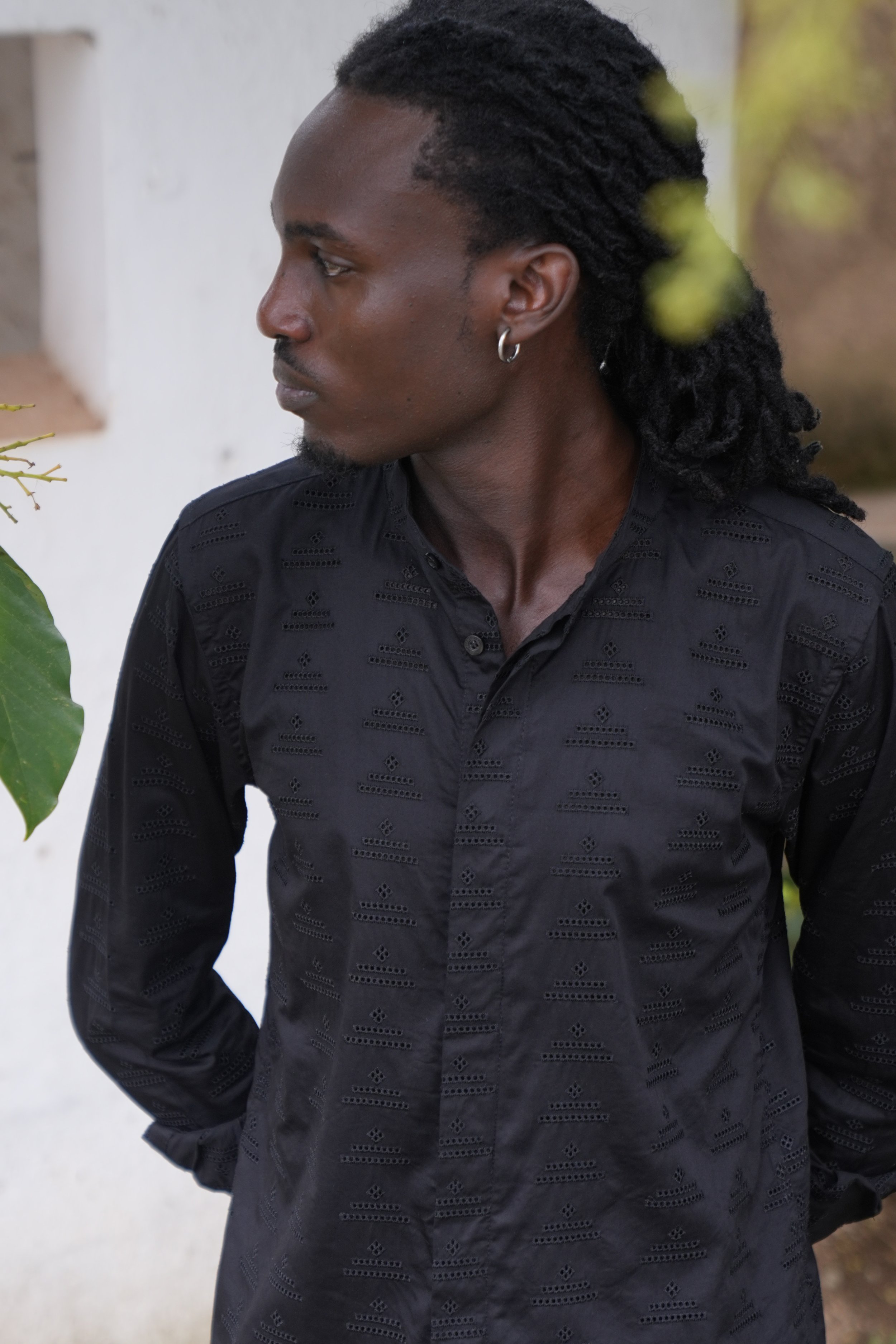 Profile view of a man with dark skin, black dreadlock hairstyle, wearing earrings and a dark patterned shirt, standing outdoors by some greenery and a white wall.