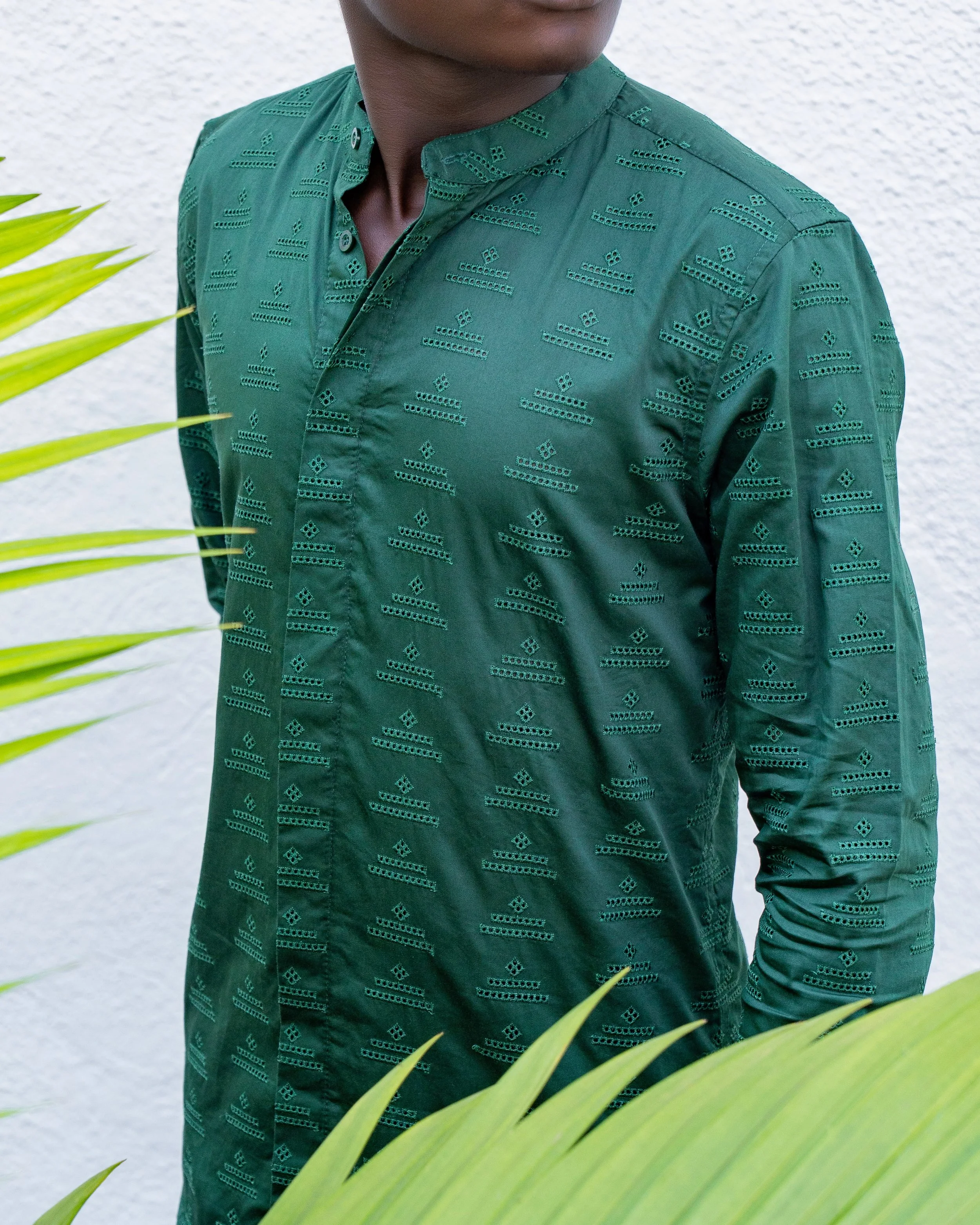 Close-up of a person wearing a green shirt with eyelet embroidery, with part of their face and shoulder visible, standing near green leaves and a white textured wall.