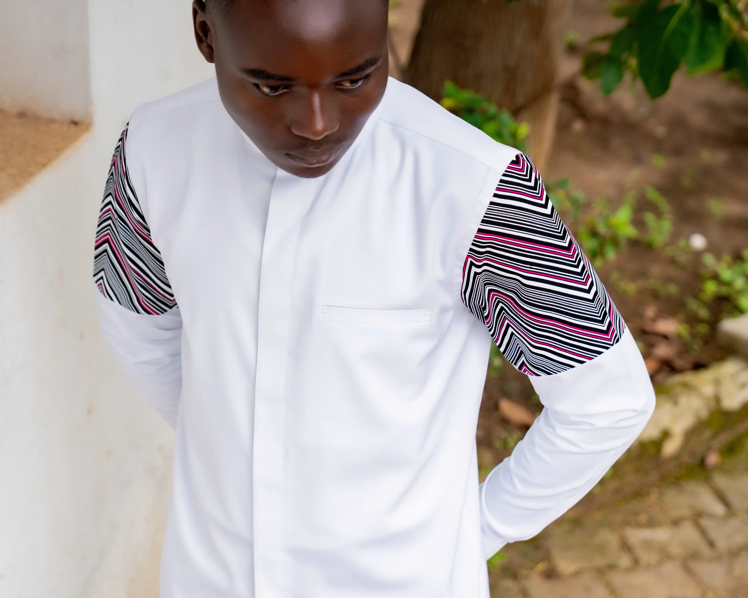 Close-up of a person wearing a white shirt with black, pink, and white patterned sleeves, standing outdoors near a tree and green foliage.