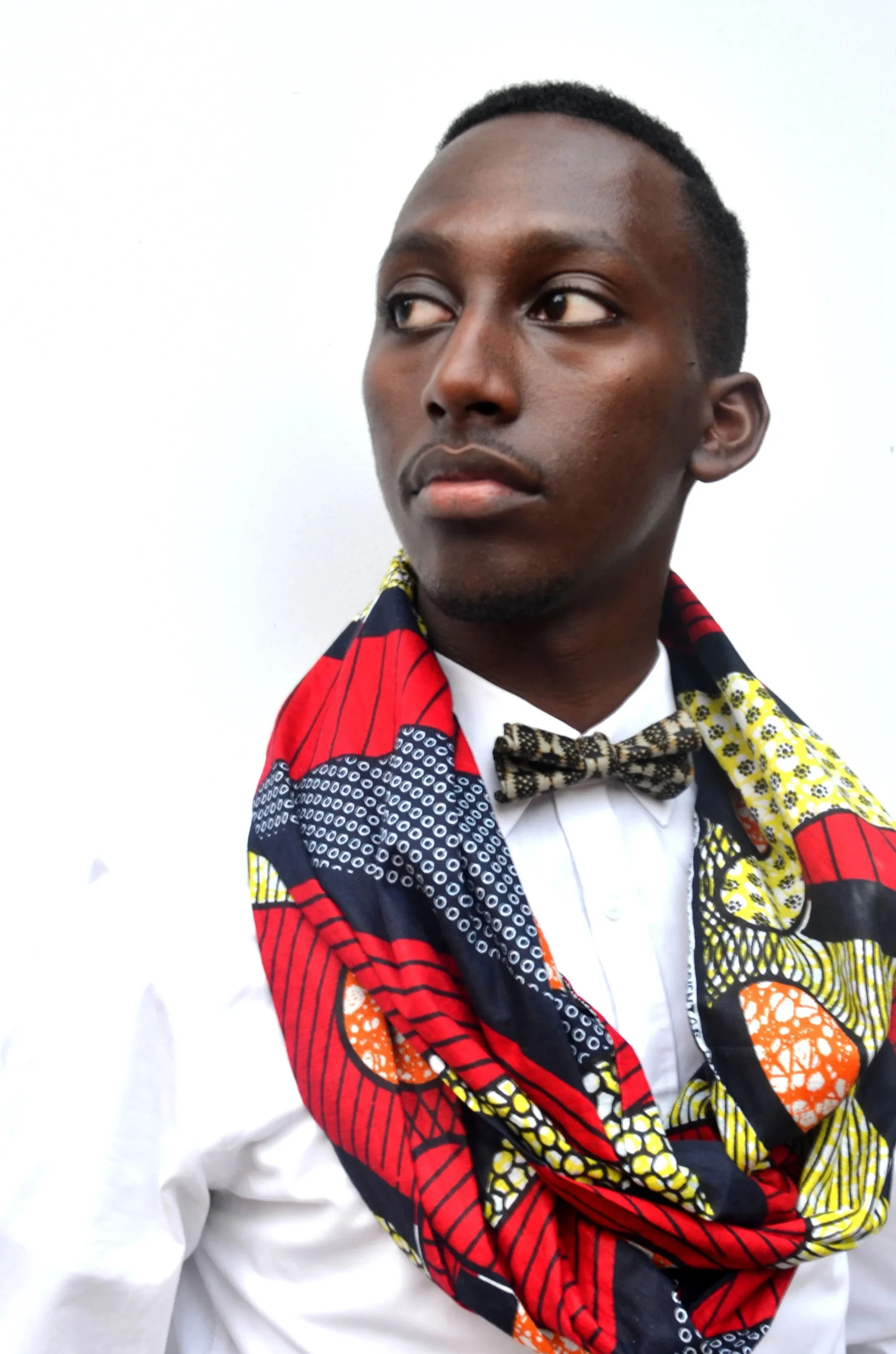 A young man in a white shirt and a colorful African print scarf, looking to the side against a plain white background.