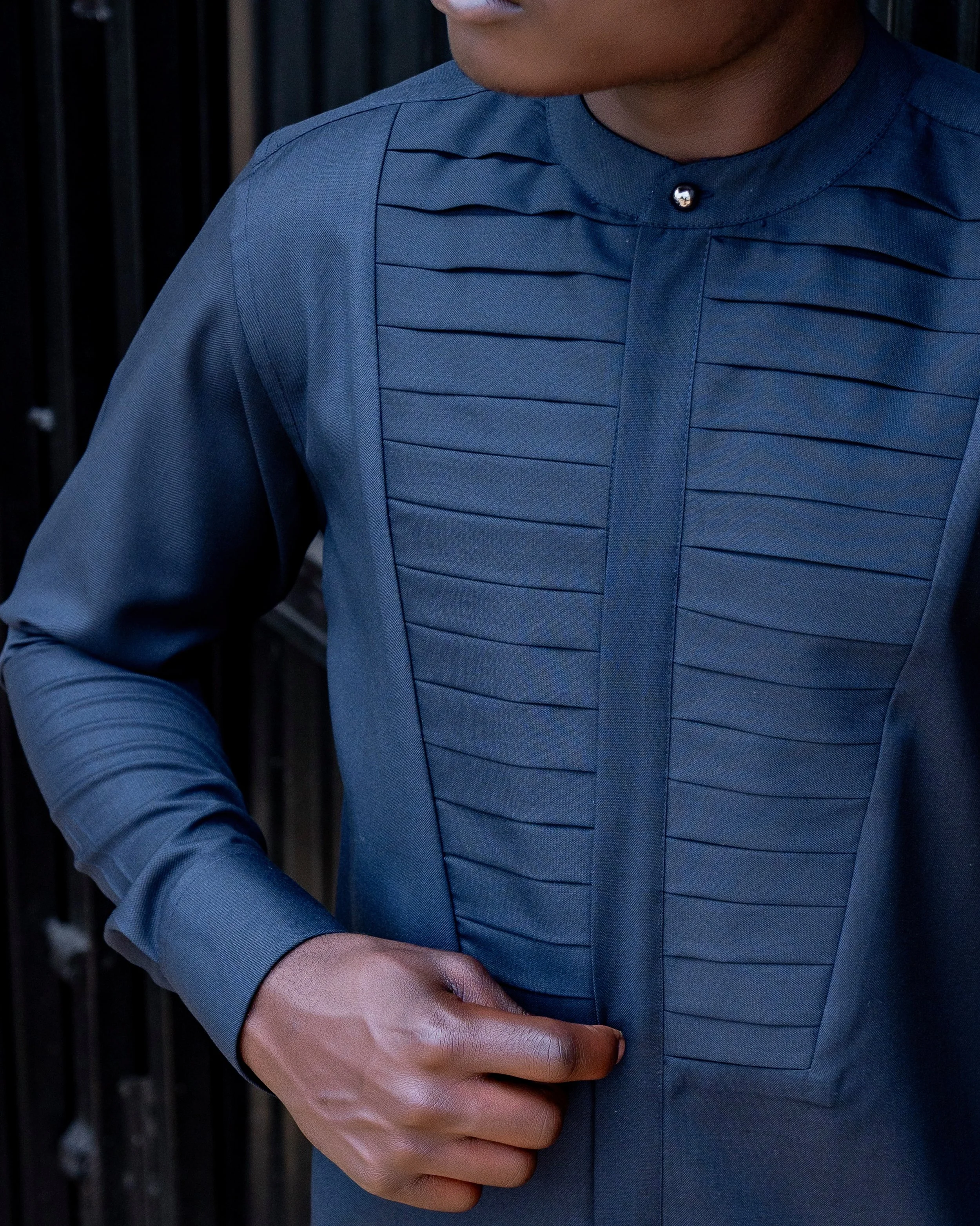 Person wearing a blue shirt with pleats on the chest, standing outdoors with a dark background, hands clasped together.