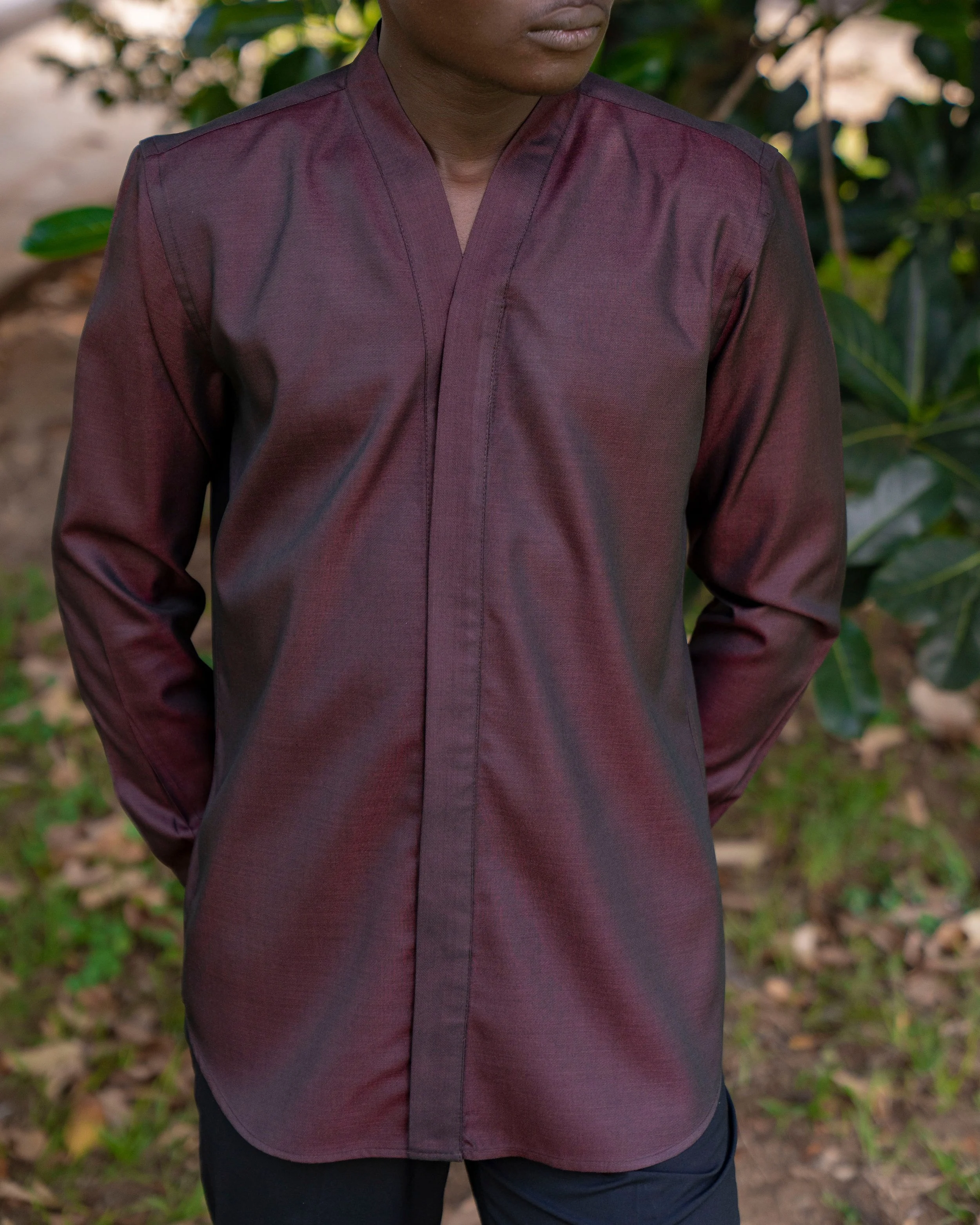 Close-up of a person wearing a dark maroon v-neck shirt with a mandarin collar, standing outdoors with green foliage in the background.