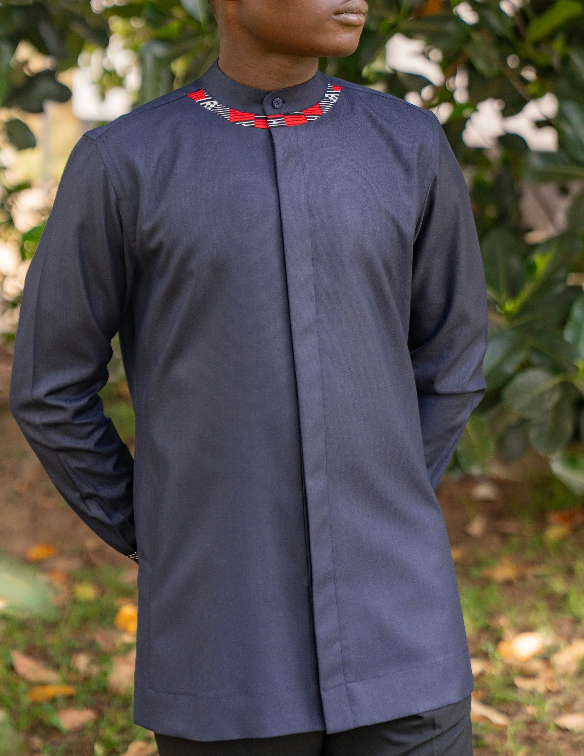 A person wearing a dark blue traditional shirt with a red, black, and white patterned collar, standing outdoors with green leaves in the background.
