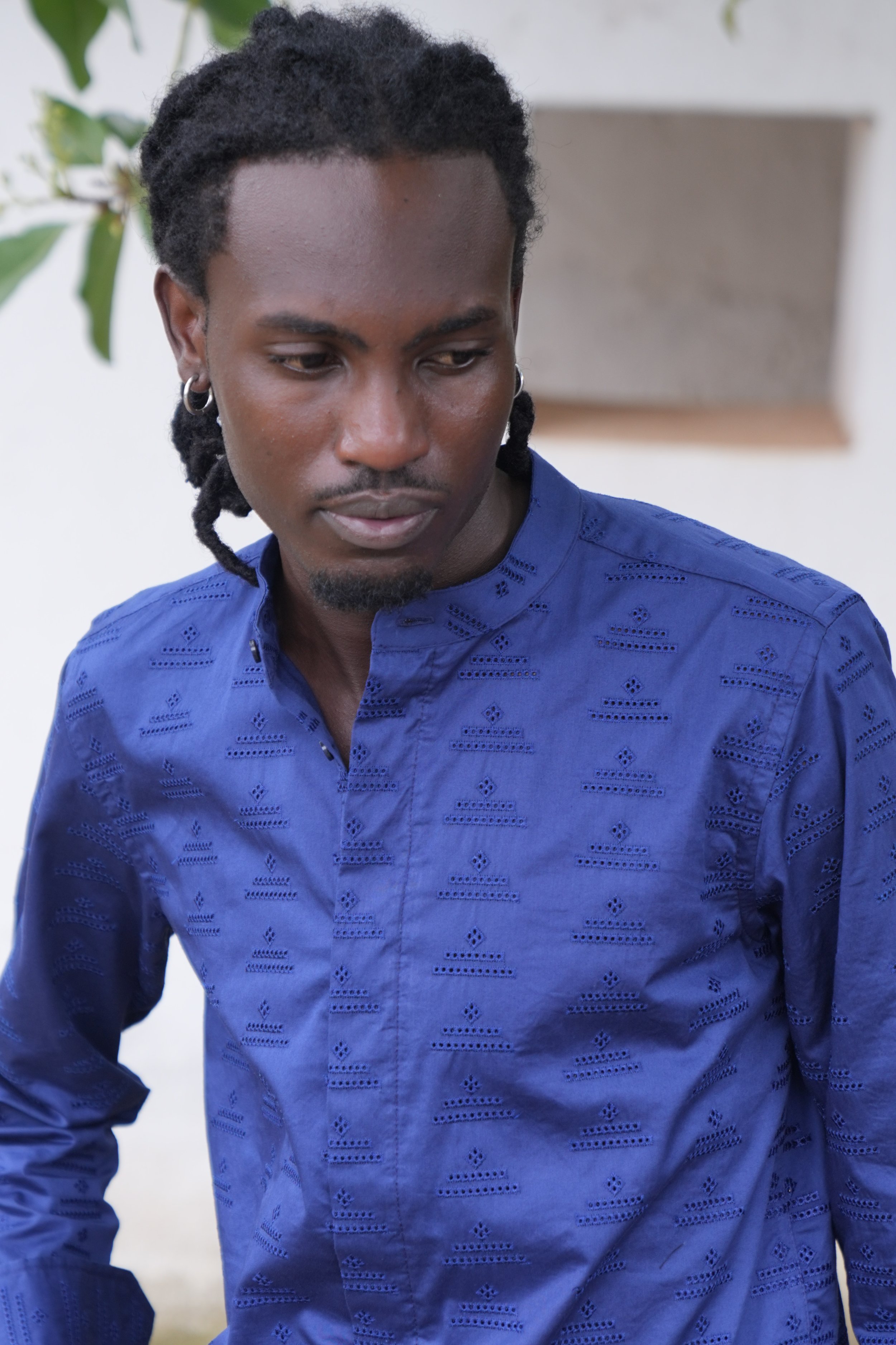 A man with dark skin and dreadlocks wearing a blue shirt with embroidery, looking down, standing in front of a white wall with a rectangular cutout.