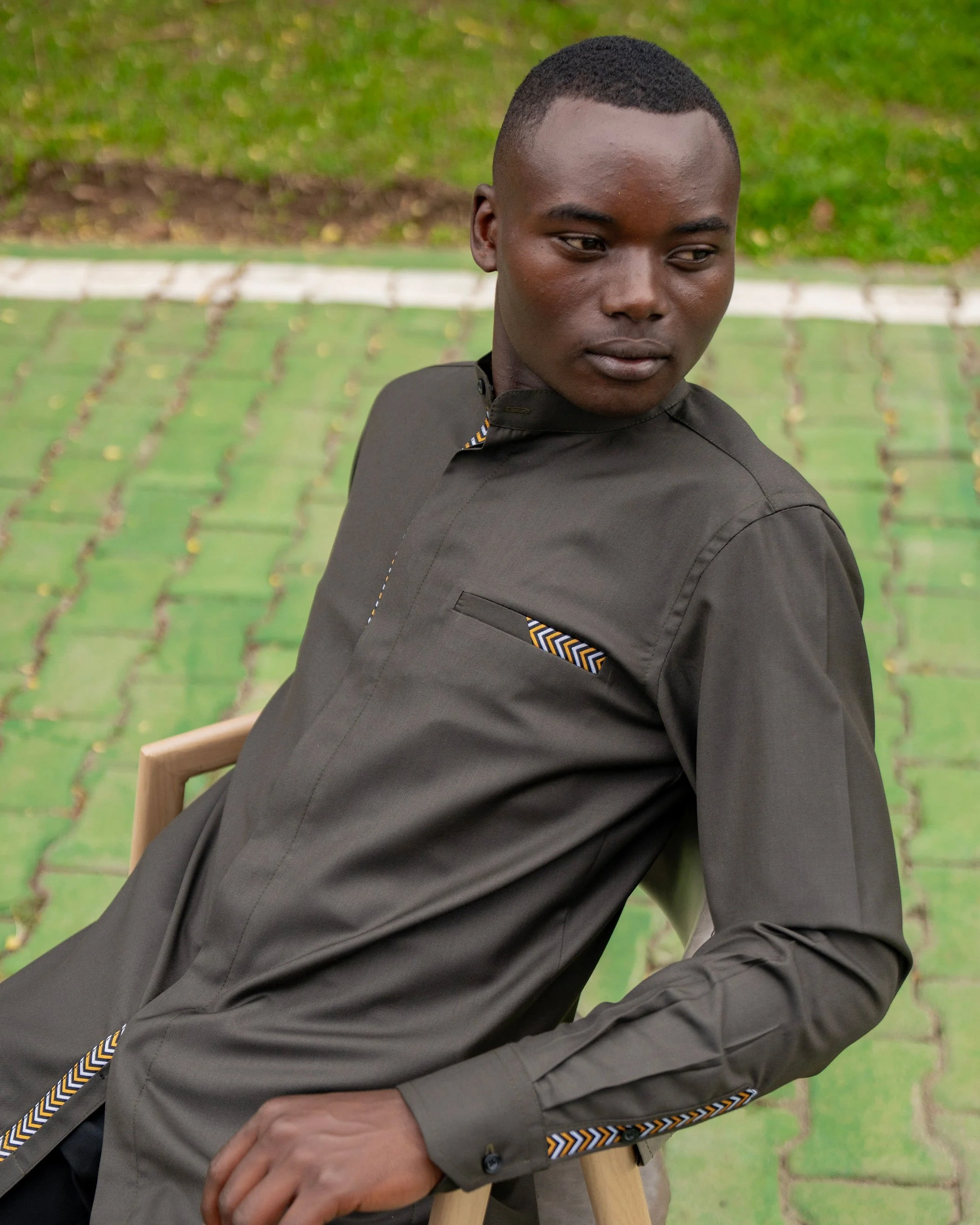 A young man with dark skin and short hair, wearing a dark gray shirt with a patterned trim, sits outdoors on a wooden chair. He is looking down and to the side with a neutral expression, in front of a backdrop of green grass and a paved path.