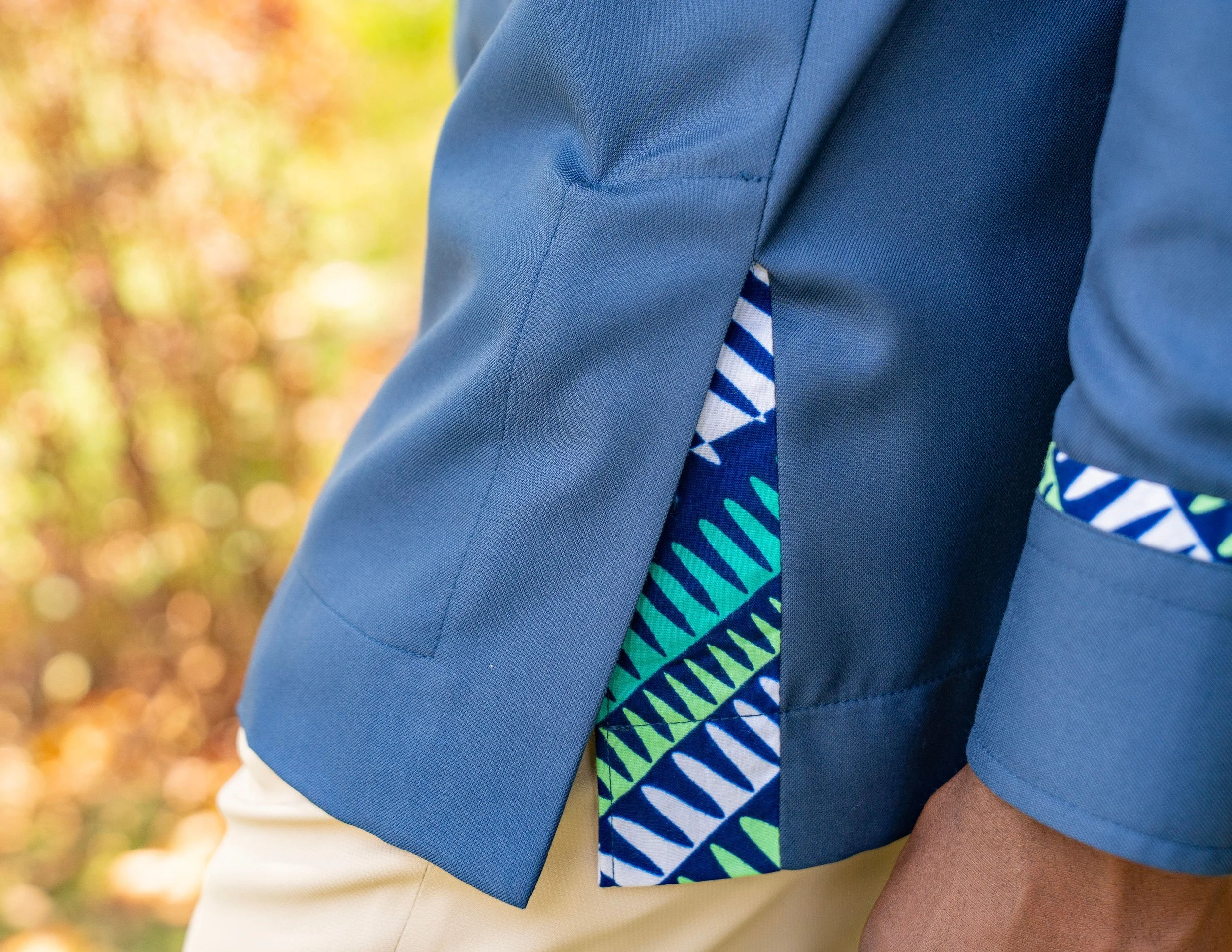 Close-up of a man wearing a blue shirt with a patterned slit detail, beige pants, and a patterned shirt. Background blurred with warm autumn colors.