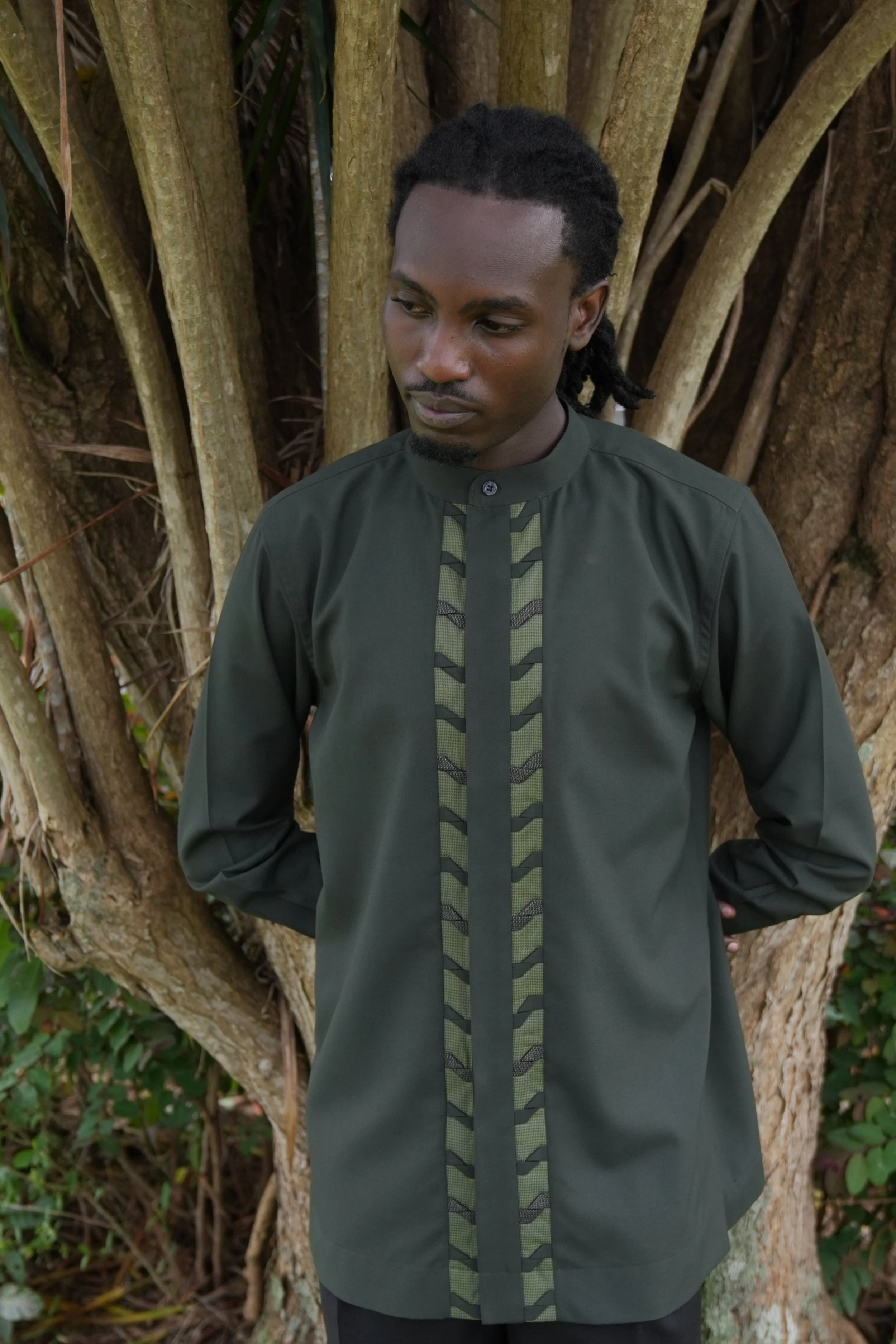 A man in traditional African attire standing outdoors in front of a large tree with textured bark and green foliage.
