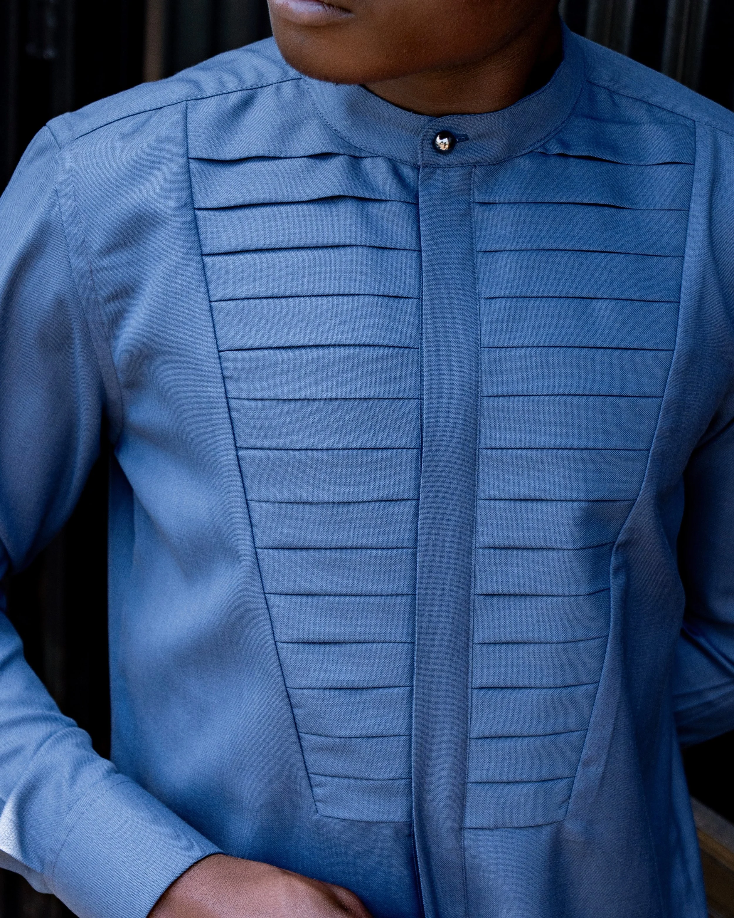 Close-up of a person wearing a blue shirt with pleated design and a mandarin collar, against a dark blurred background.