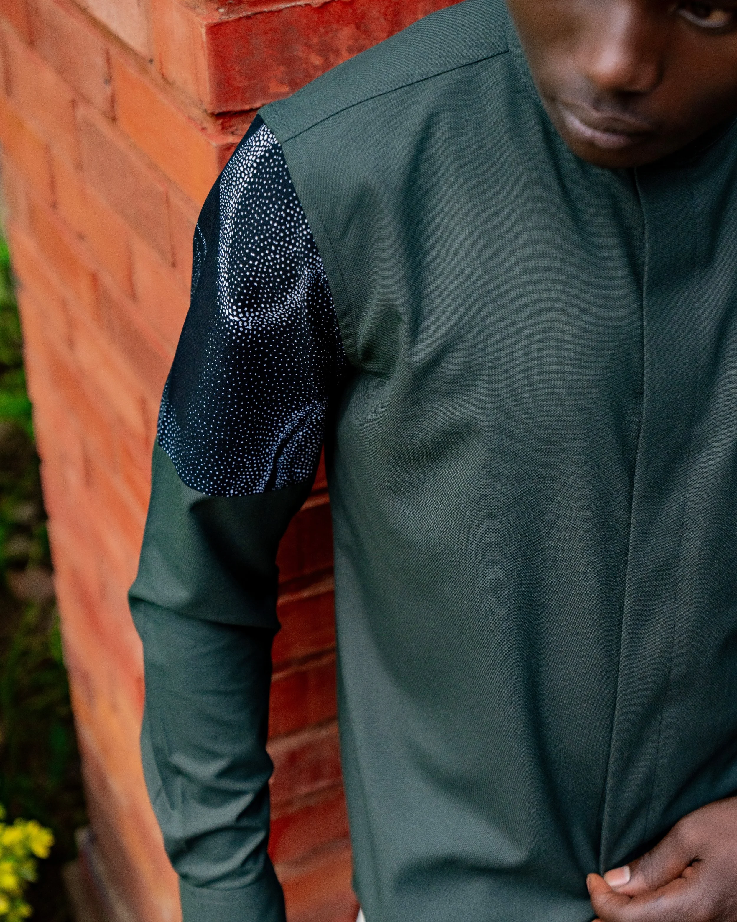 Close-up of a person wearing a dark green shirt with a dotted pattern on the shoulder, standing in front of a red brick wall.