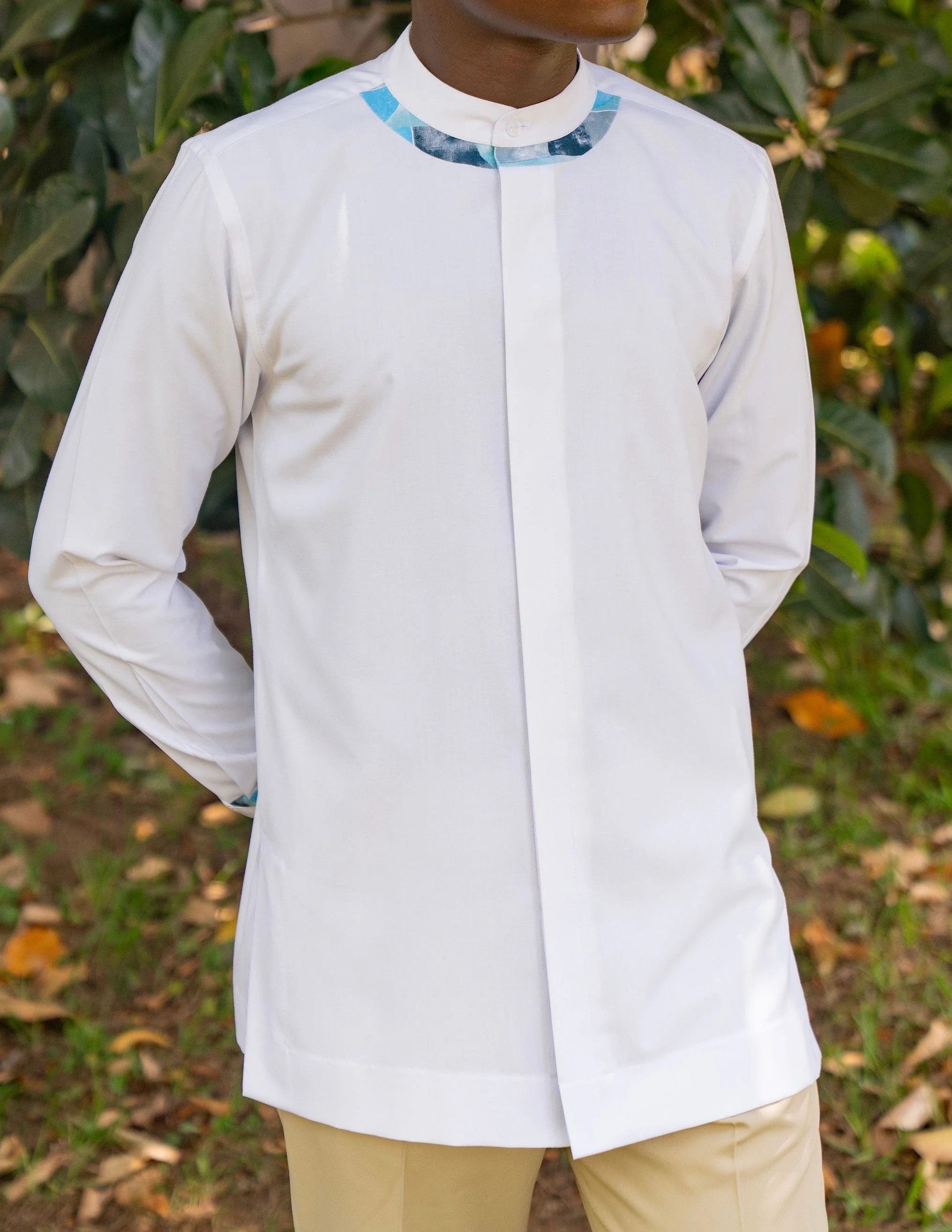 Person wearing a white shirt with a detailed collar, beige pants, standing outdoors with green foliage and fallen leaves in the background.