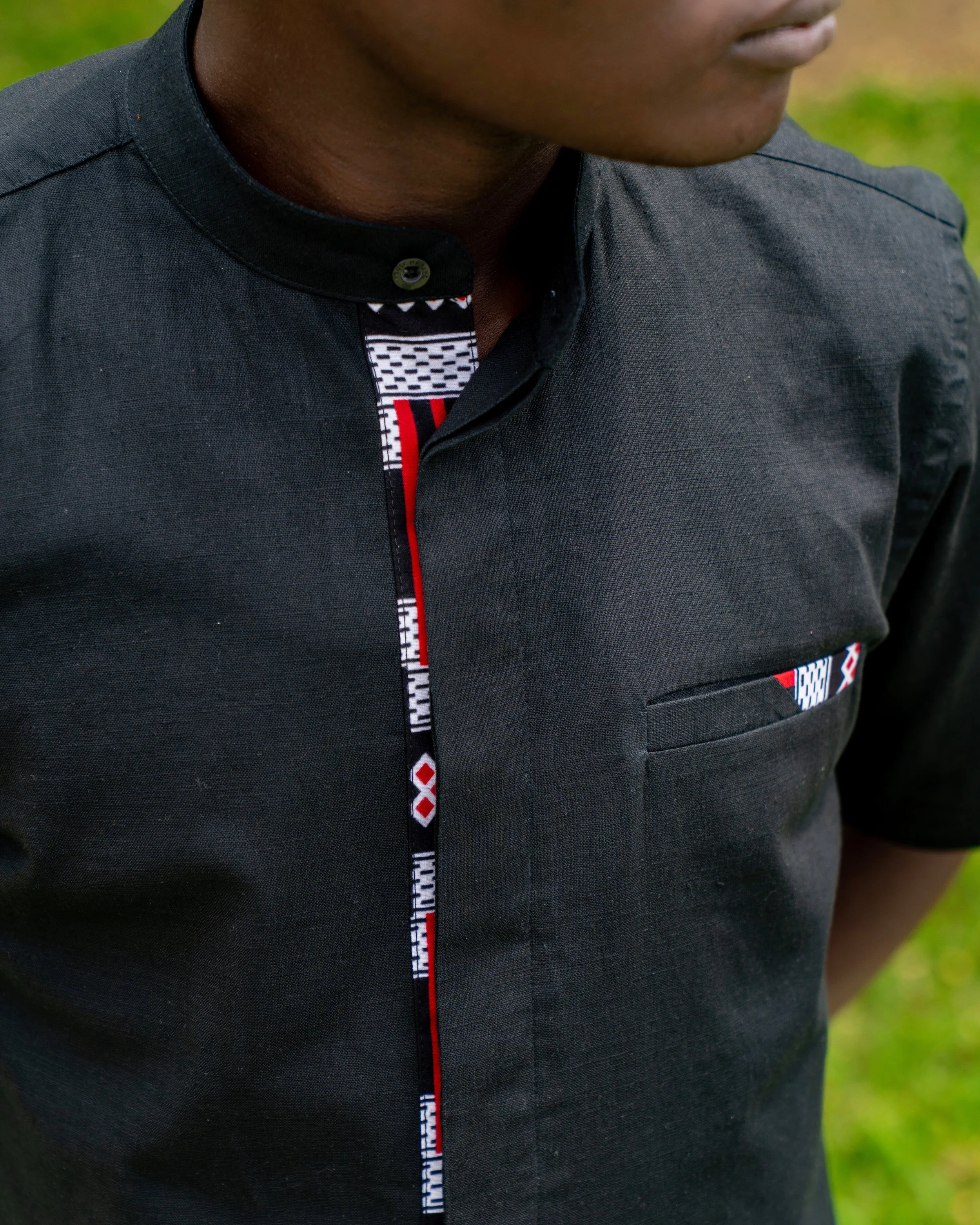 Close-up of a person wearing a black shirt with a patterned collar and button, standing outdoors with green grass blurred in the background.