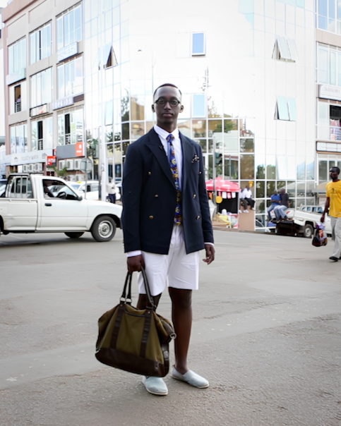 Young man standing on city street, wearing a dark blazer, white shorts, and carrying a large brown bag.