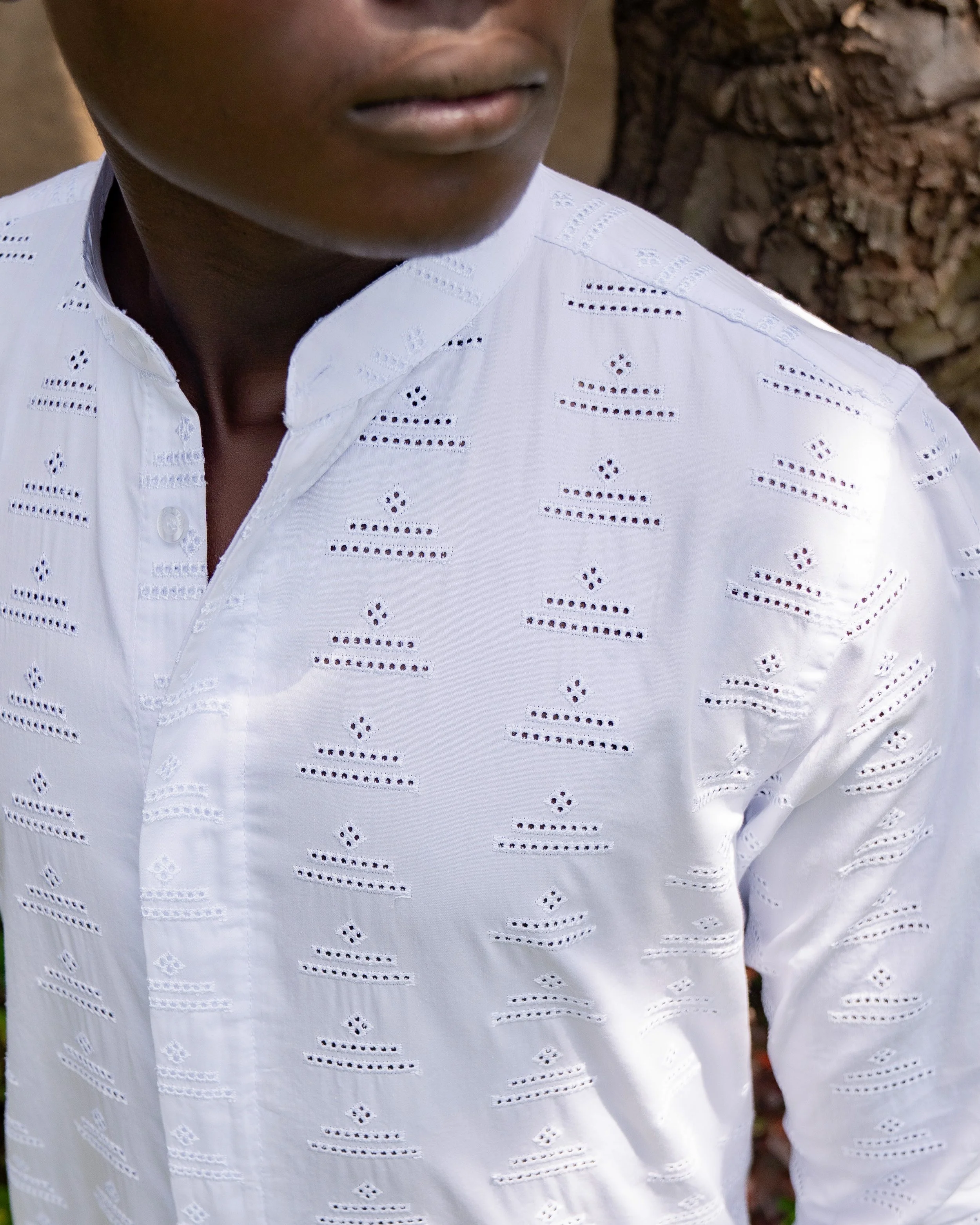 Close-up of a person's face and upper torso, wearing a white embroidered shirt with cutout patterns, standing outdoors near a tree trunk.
