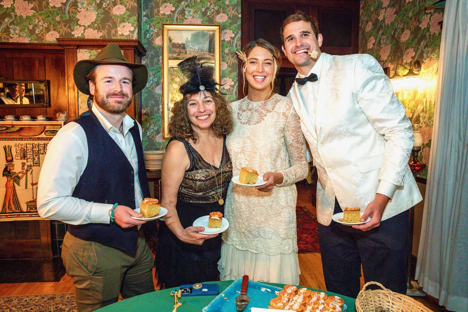 A group of four people in festive attire holds plates of cake, smiling in a decorated room with floral wallpaper.
