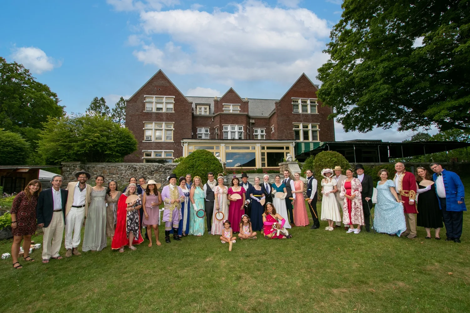 A large group of people in vintage attire poses on a lawn in front of a grand brick building under a blue sky.