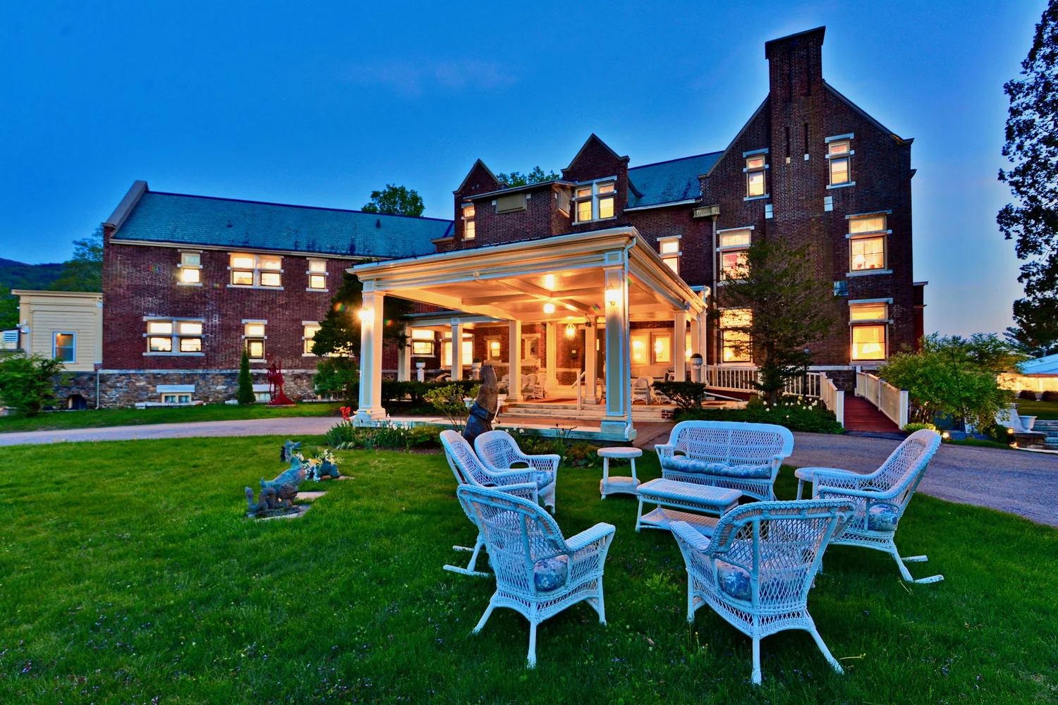 A charming brick building with a porch, surrounded by green grass and white wicker furniture, illuminated at dusk.