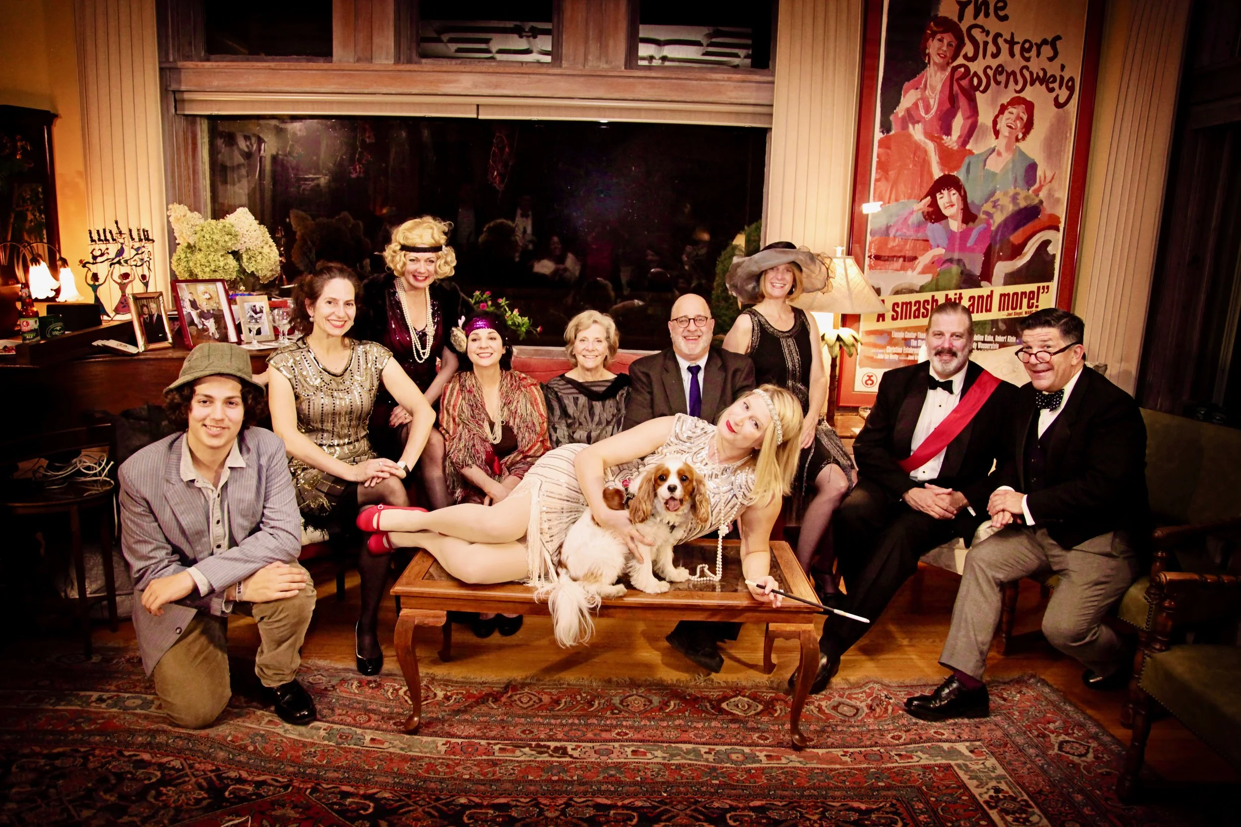 A group of people dressed in vintage attire poses together in a cozy room, featuring a dog on a table and a theatrical poster in the background.