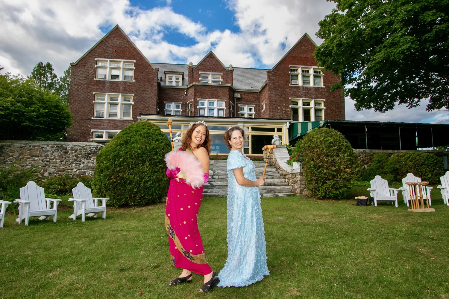Two women pose back-to-back in a garden, dressed in vibrant gowns, with a large brick building in the background.