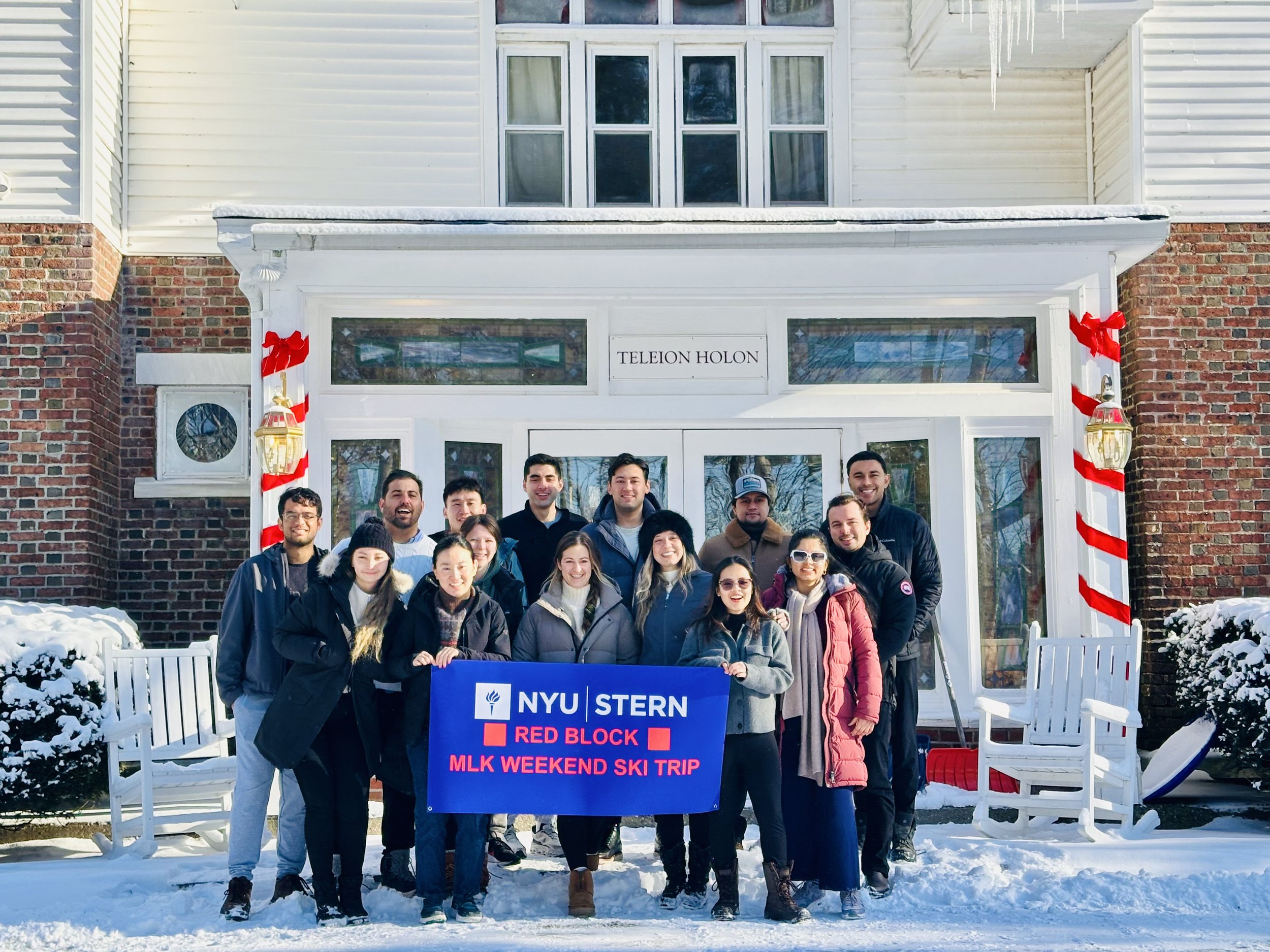 A group of people poses in front of a snowy building, holding a banner for an NYU Stern ski trip. Decorations and snow are visible.
