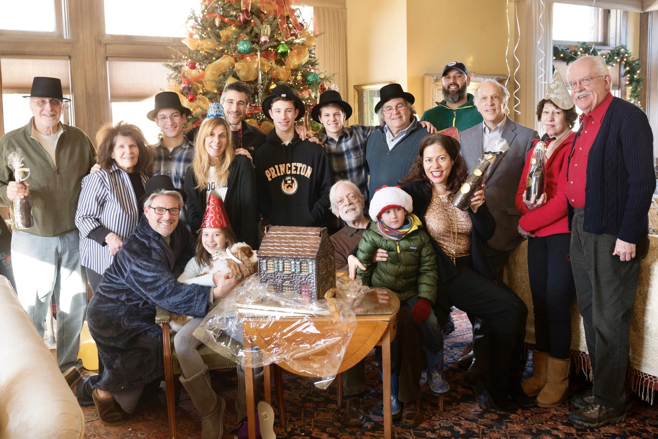 A large family gathering in festive attire, posing by a decorated Christmas tree with a gingerbread house on a table.