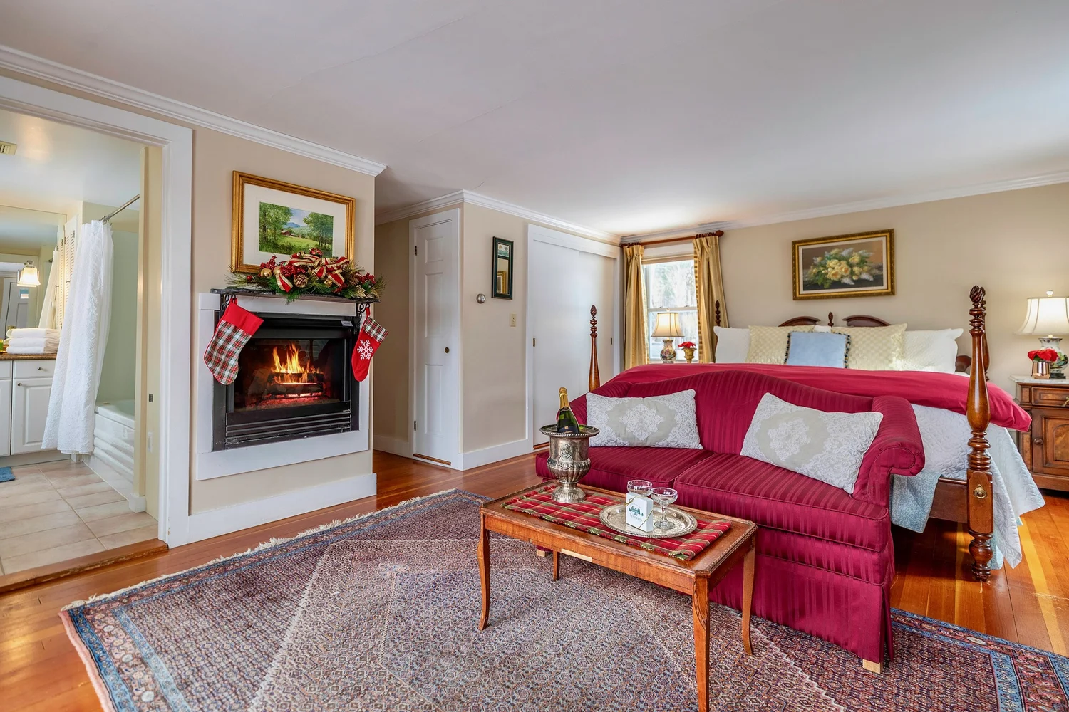 Cozy bedroom with a fireplace, red sofa, and festive stockings. Features a bed with decorative pillows and a small table with drinks.
