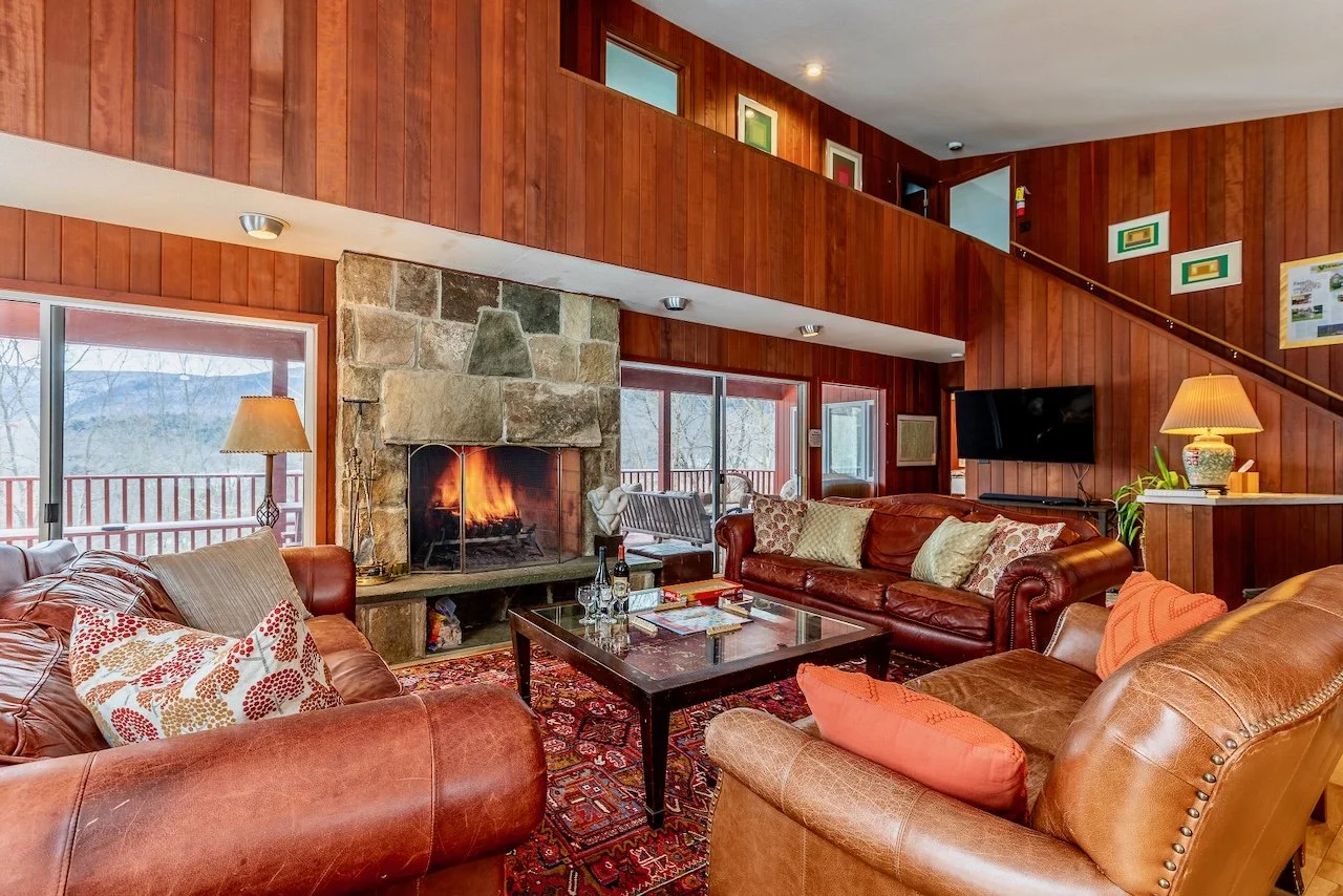 Cozy living room with a stone fireplace, leather sofas, and large windows showcasing a scenic view. Warm wood paneling and decorative accents.